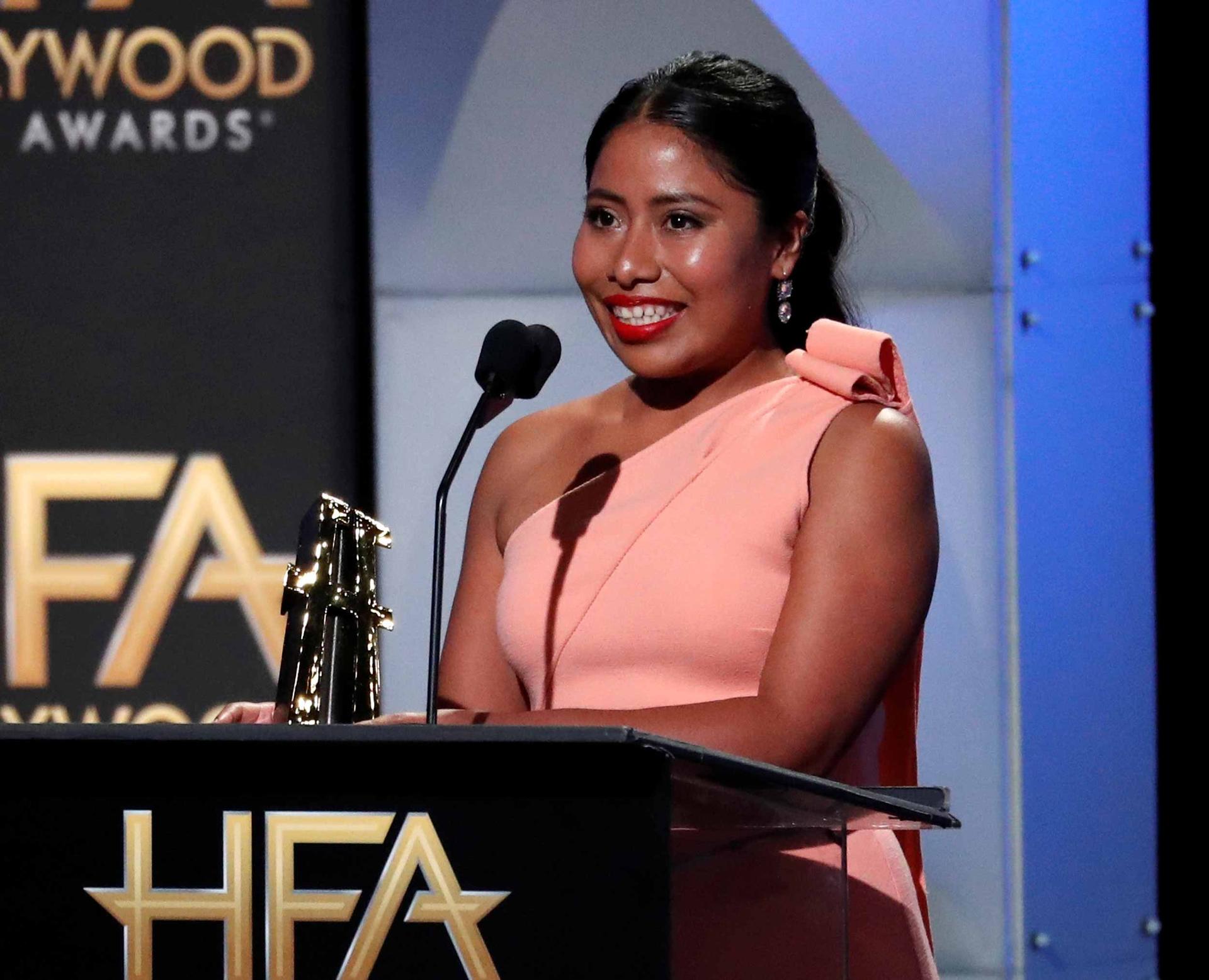Yalitza Aparicio stands behind a podium holding the New Hollywood Award