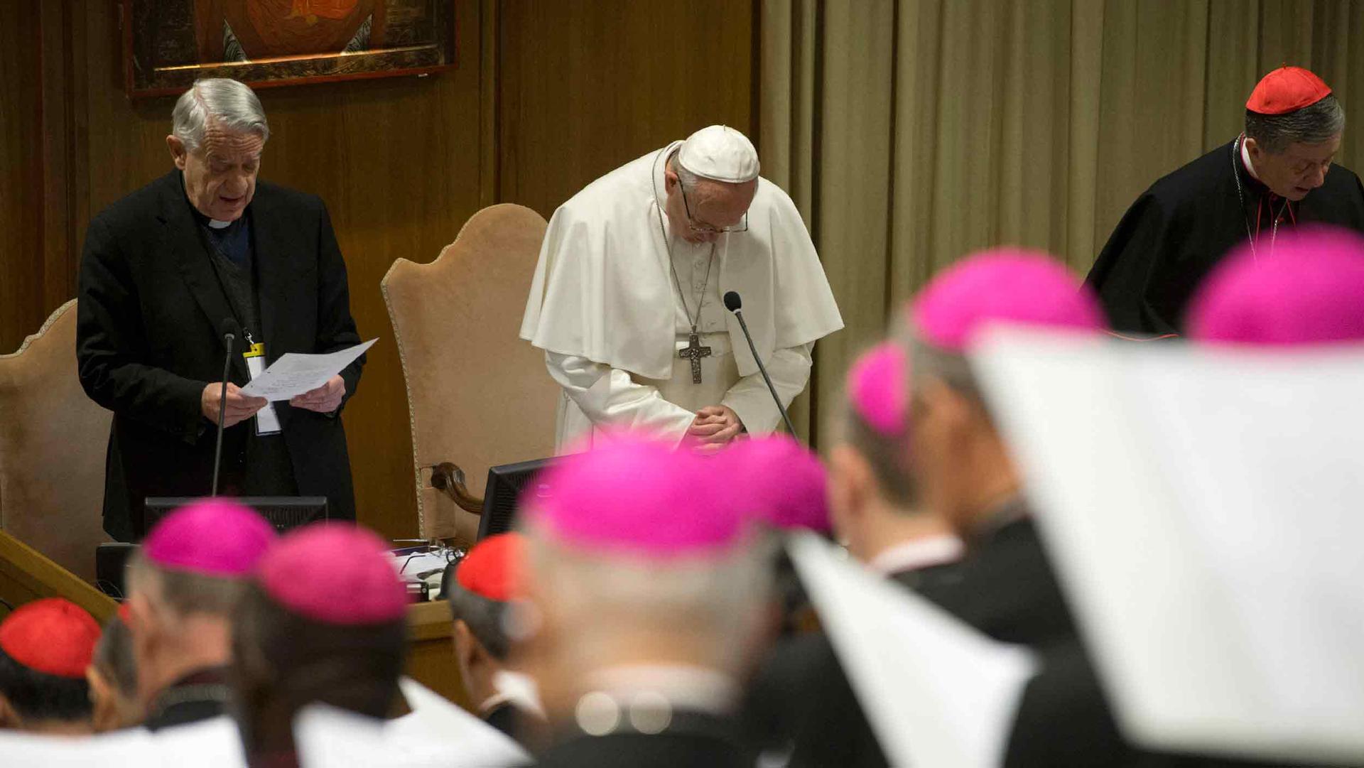 Pope Francis bows his head in prayer. Around him are other members of the clergy wearing red caps