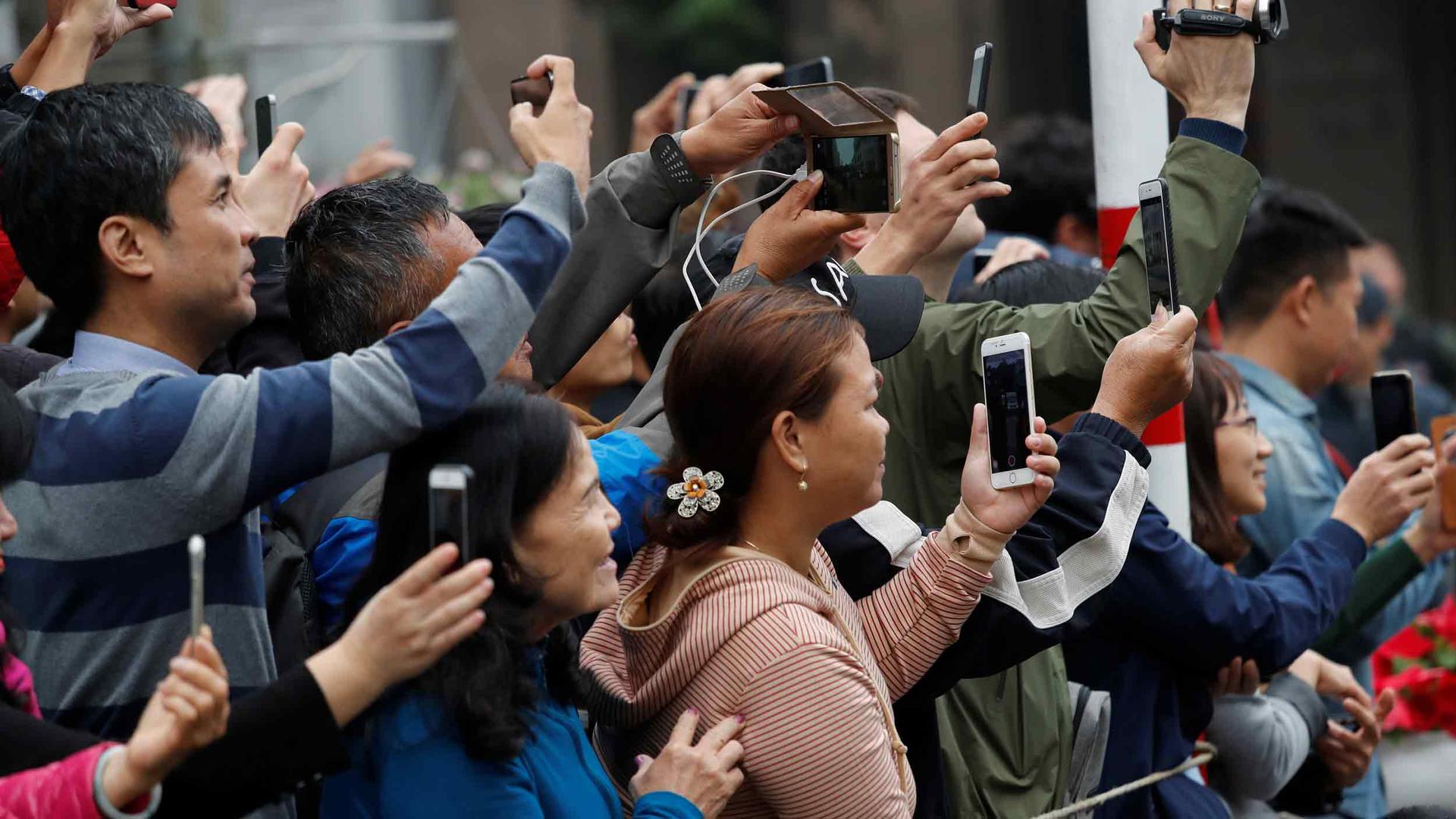A crowd of people hold phones and cameras to take pictures as the motorcade carrying Kim Jong-un passes by