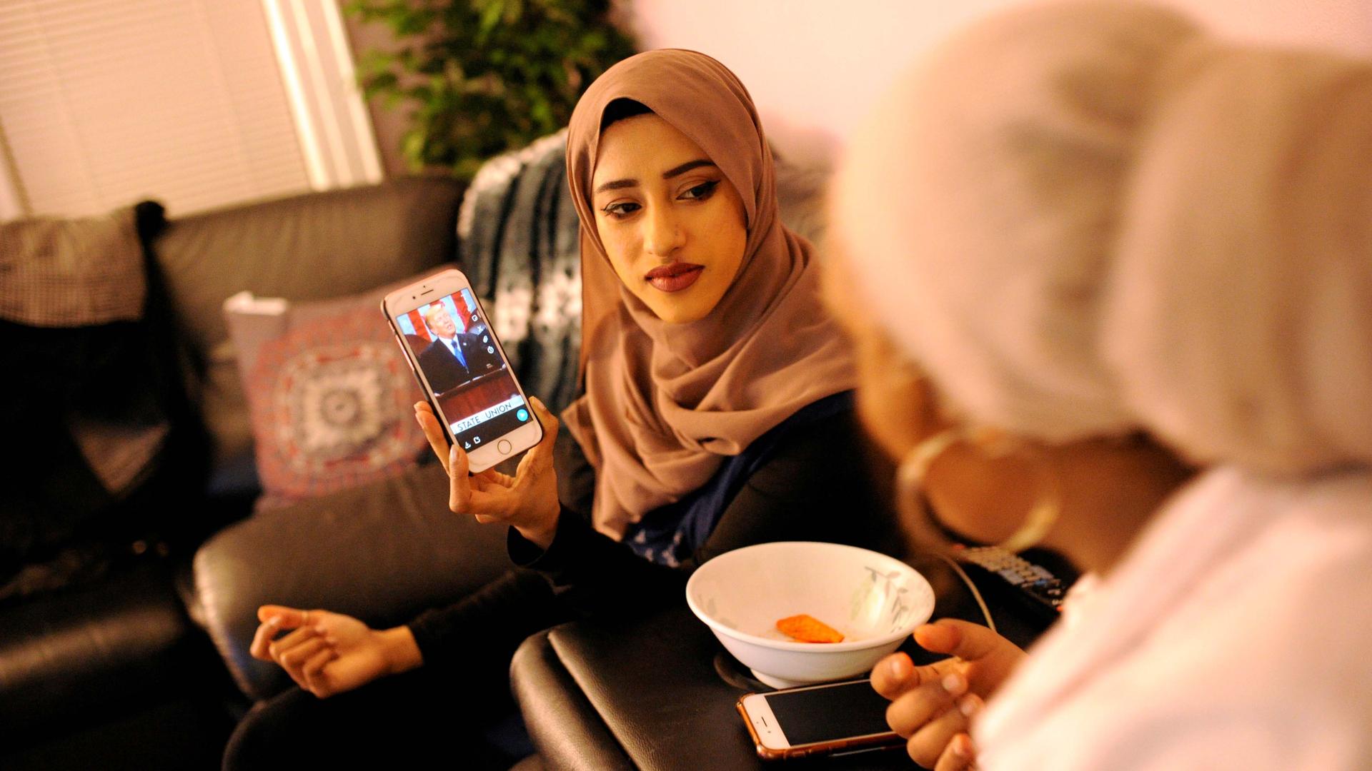 A young woman in a hijab shows her phone screen to another young woman whose back is turned to the camera. On the phone is Donald Trump. 