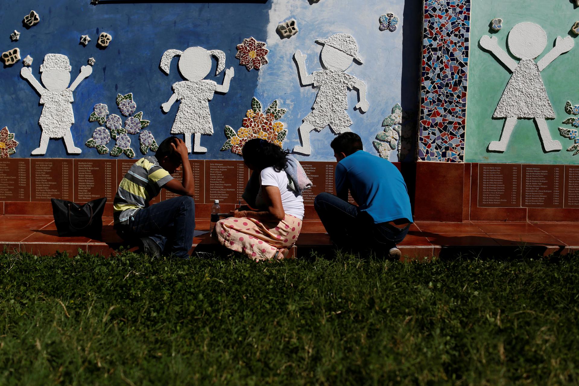 three people kneel near a wall with silhouettes of children