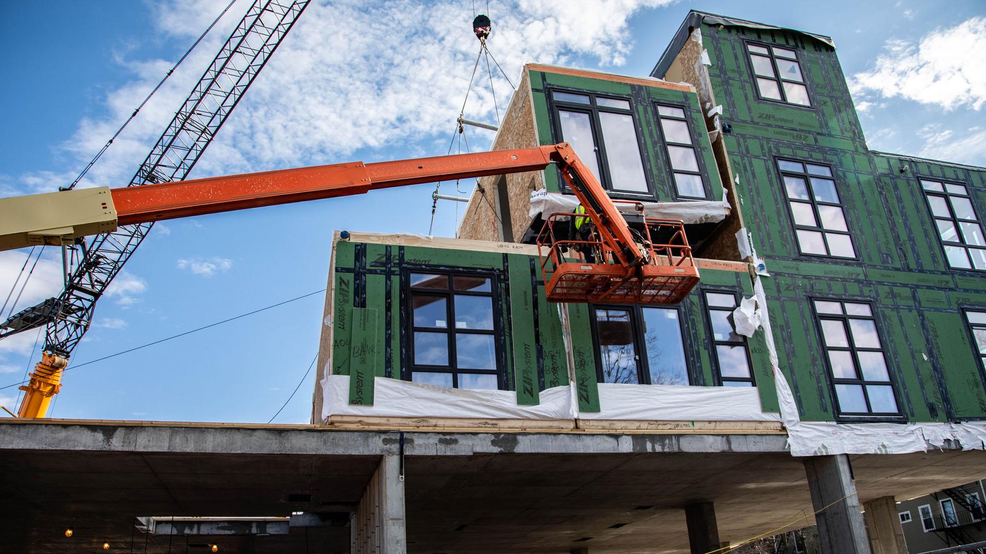 A 65 by 13 foot modular unit is shown with a green insulated side and windows, dangling from a crane.