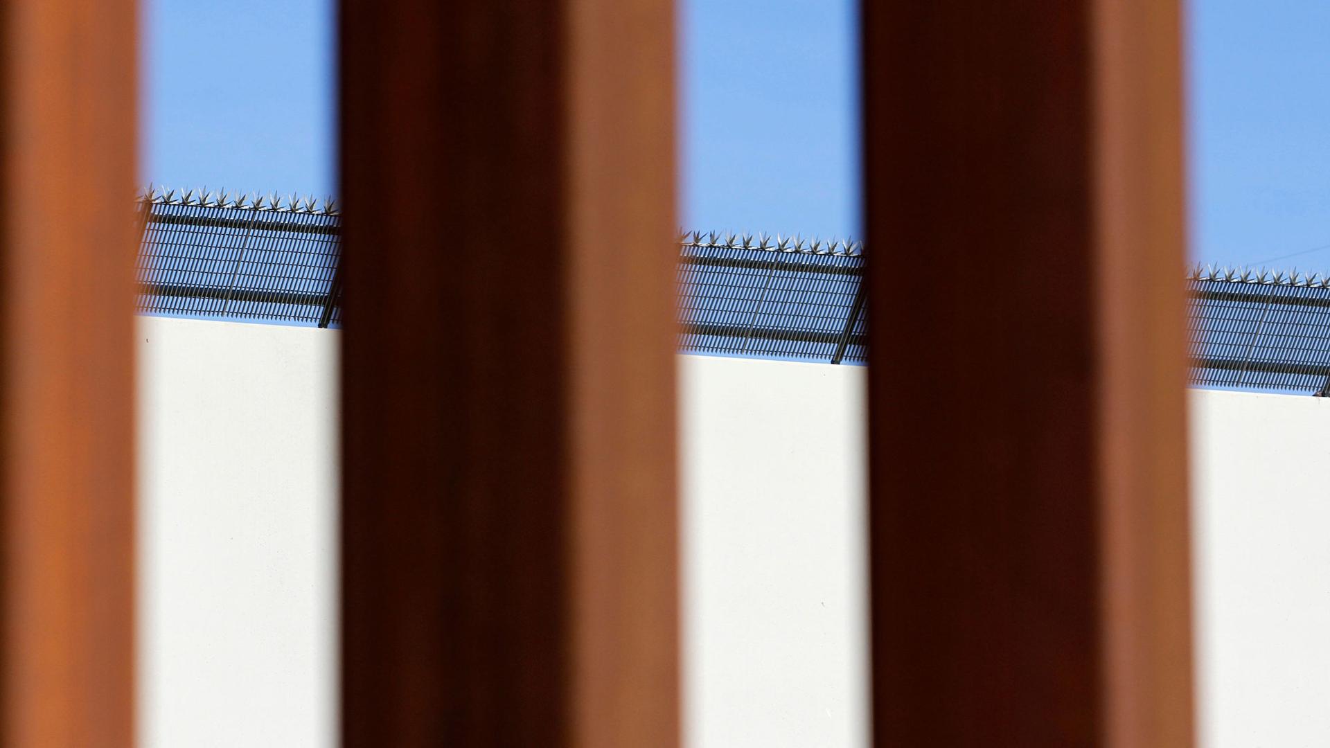 A prototype border wall with large metal spikes on top is seen through the metal slates of the border fence between Mexico and the United States.