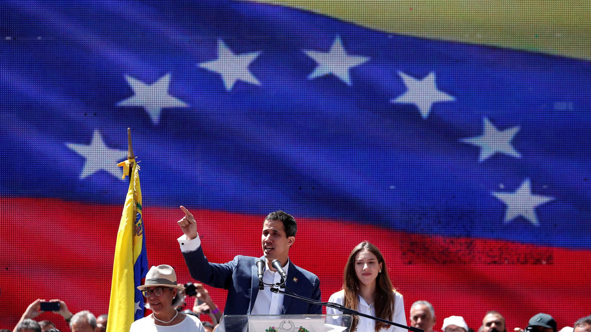 Opposition leader Juan Guaido is shown standing at a podium with a large Venezuelan flag projected behind him.