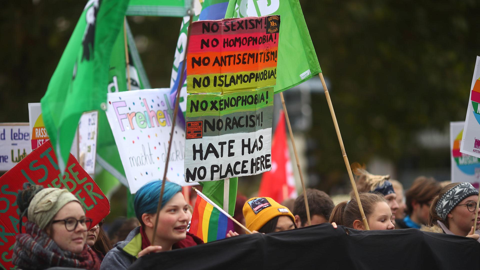 People are shown protesting against hate carrying flags and a brightly colored placard.