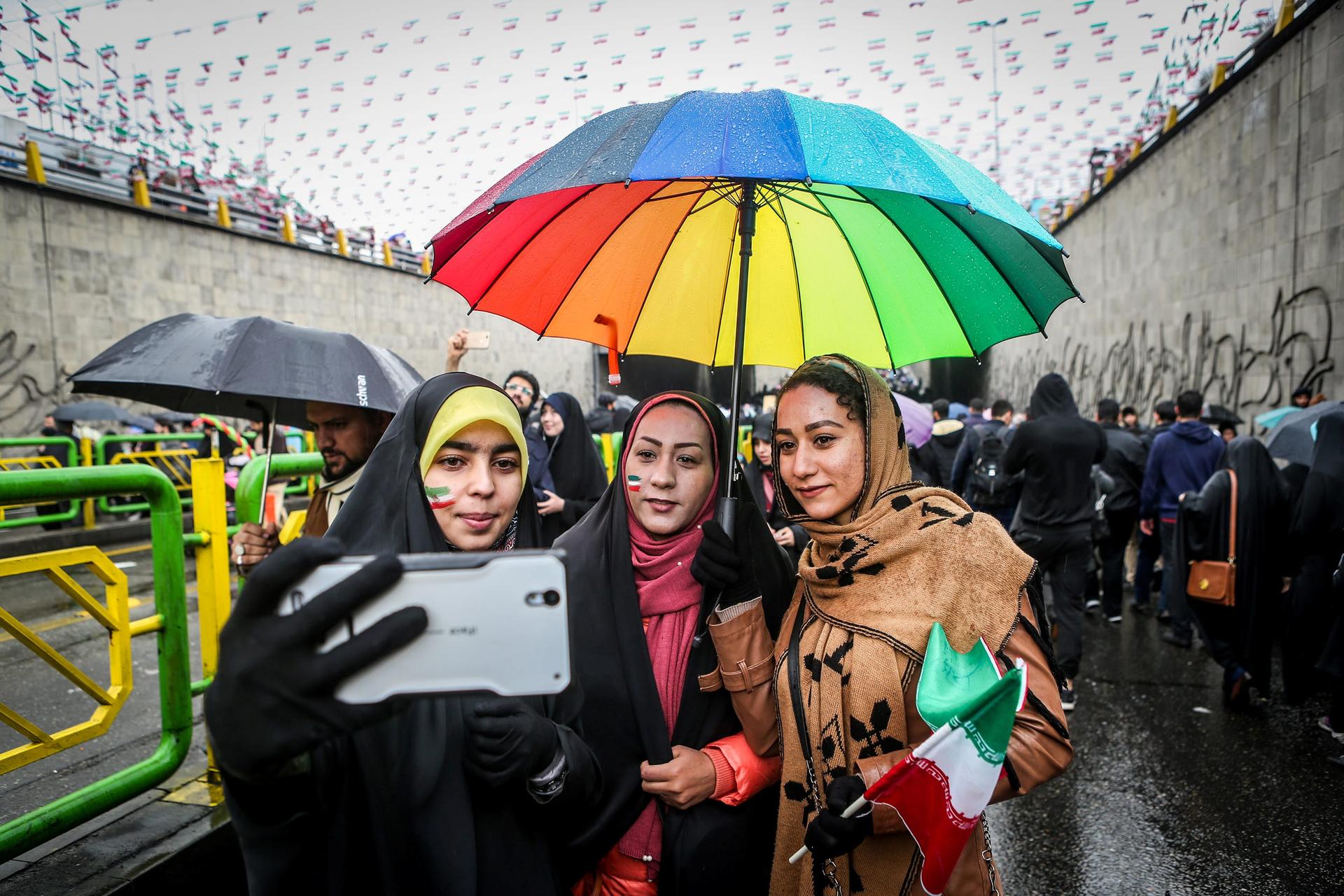 three women take a selfie with a rainbow umbrella covering them from the rain 