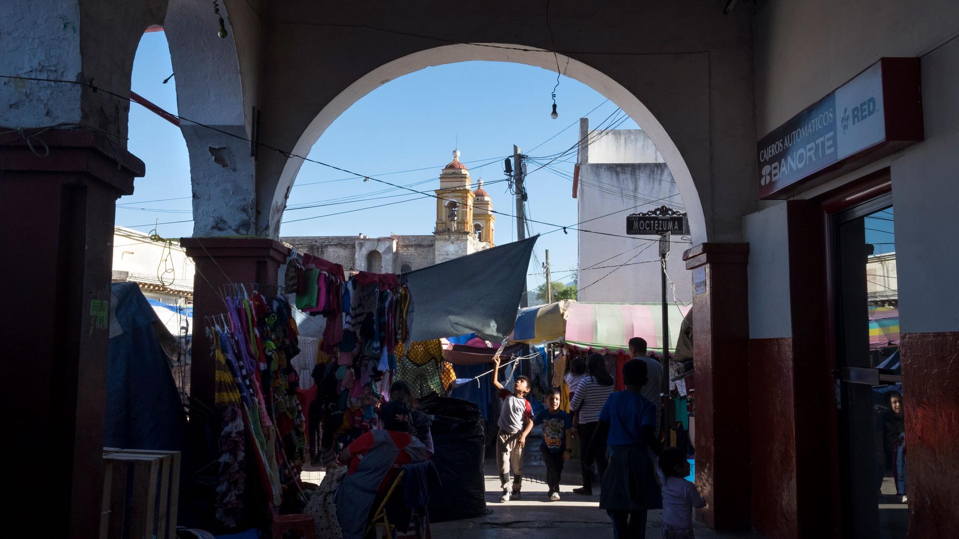 People are shown walking around at Tlaxiaco’s market, with the steeples of the Santa Maria de la Asunción church in the distance.