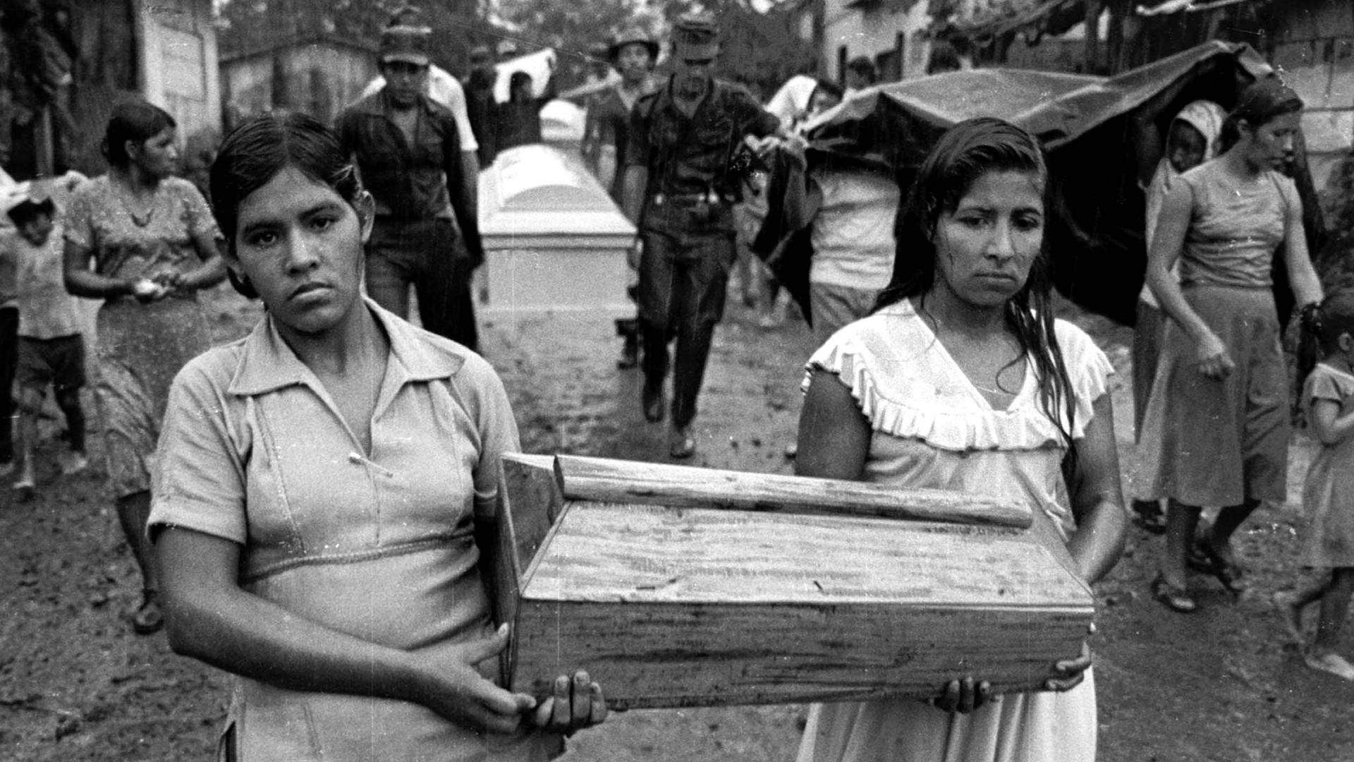 Two young women carry a small coffin between them. Black and white photo.