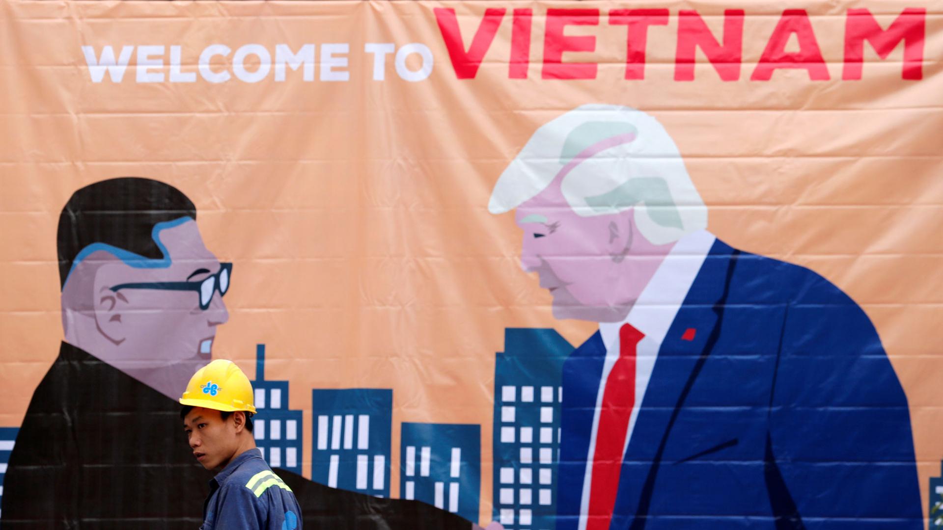 A man is shown wearing a construction hard hat walking past a banner illustration depicting North Korean leader Kim Jong-un and US President Donald Trump