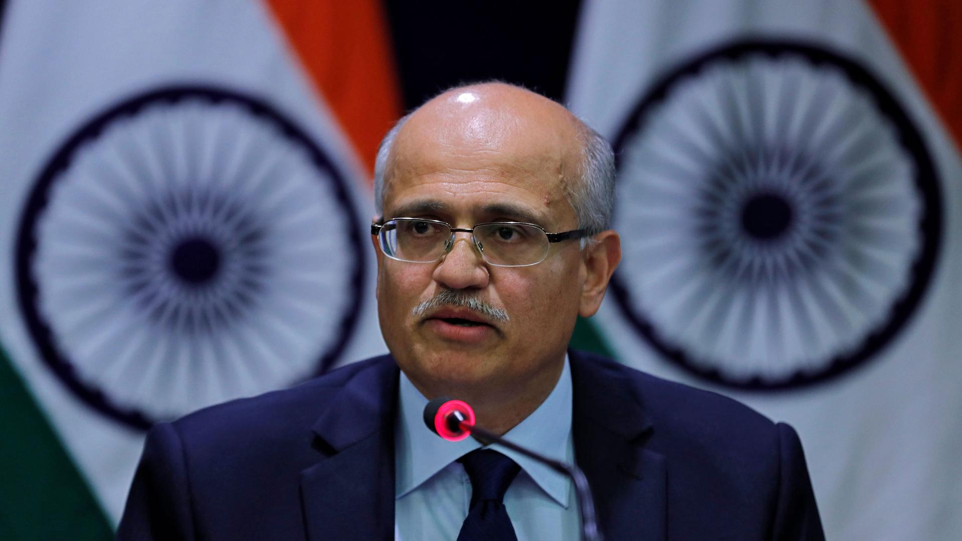 India's Foreign Secretary Vijay Gokhale is shown wearing a blue suit and speaking into a microphone with Indian flags behind him.