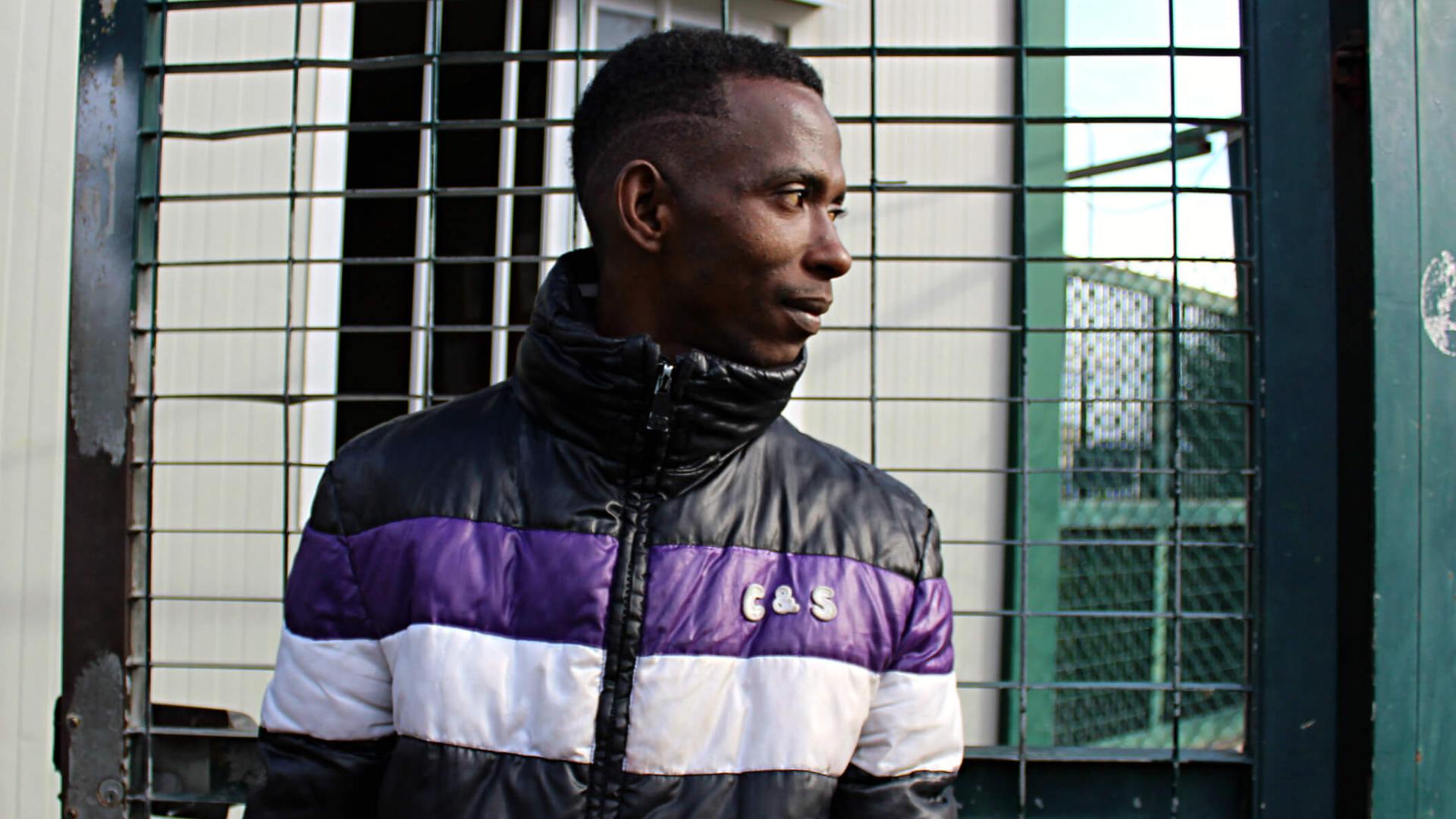A young African man wearing a purple and white jacket stands and looks to the left. 