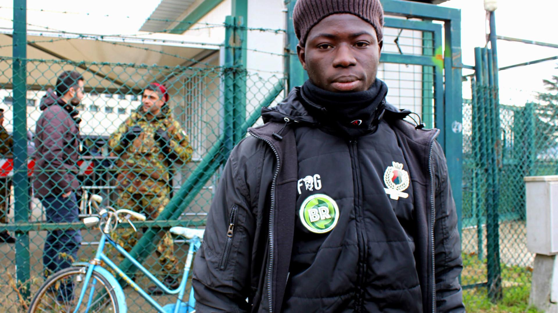 A young African man wears a green hat and a black jacket near a fence.