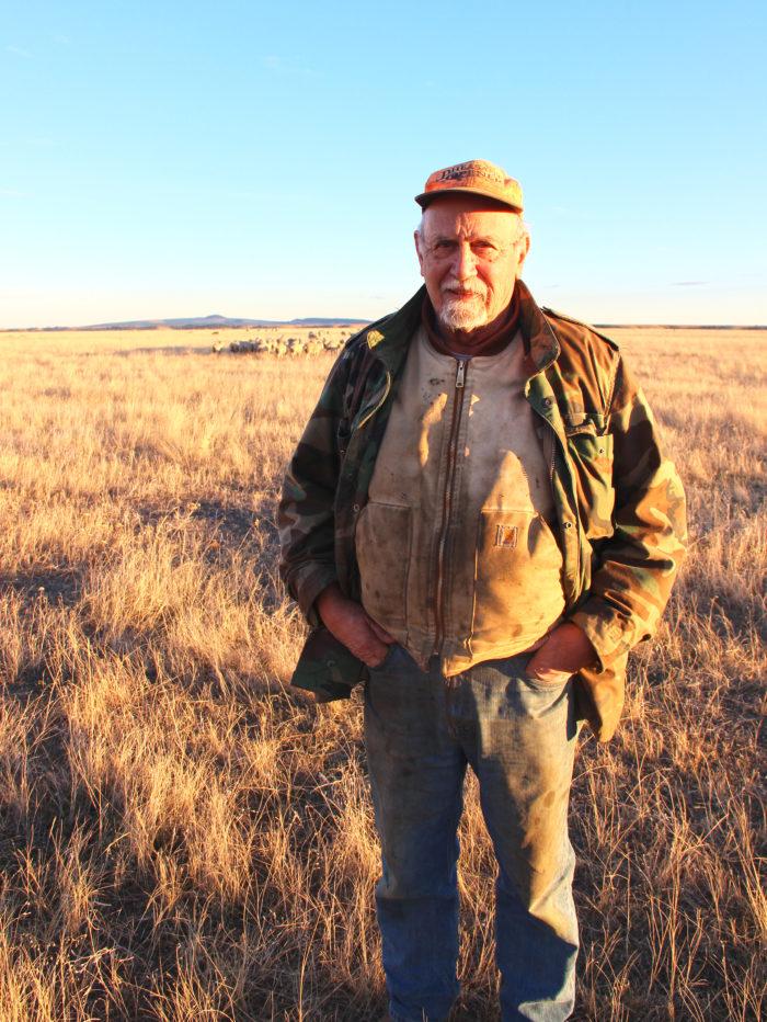 A white man wearing jeans, jacket and hat stands in yellow fields.