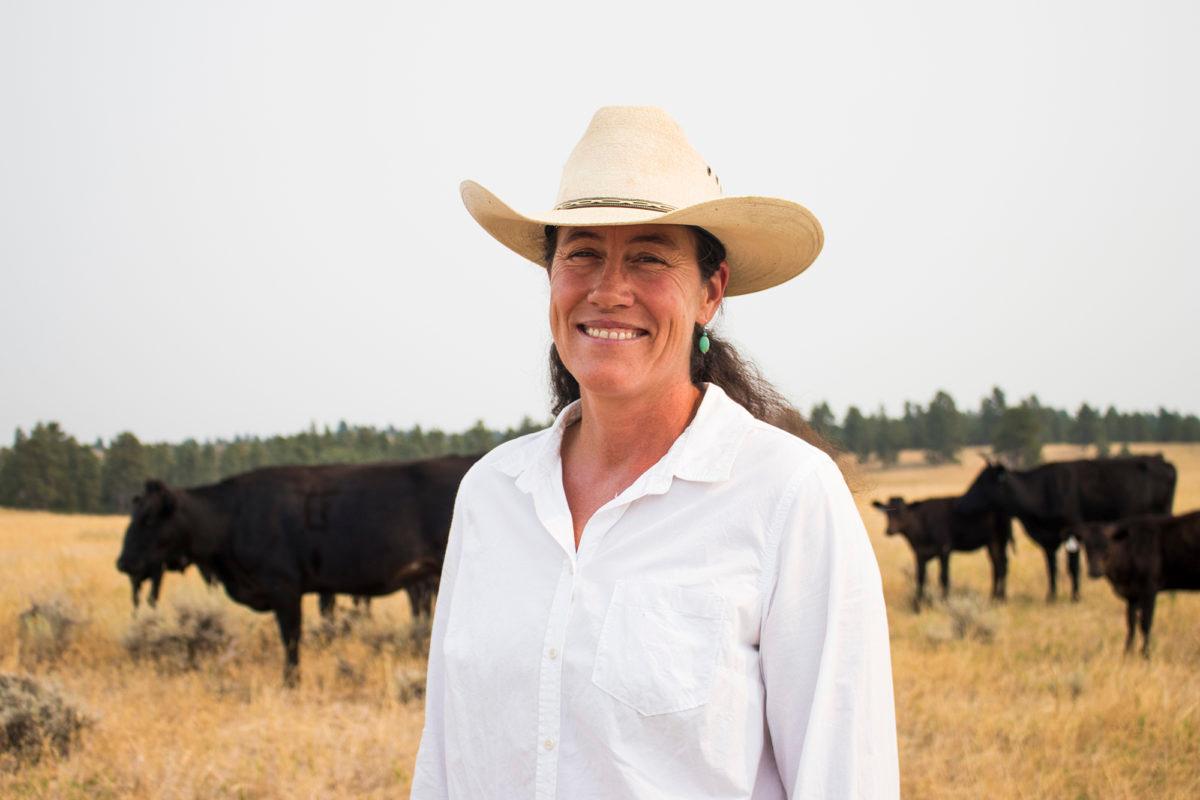 A white woman wearing a white shirt and a cowgirl hat stands in a field in front of cows. 