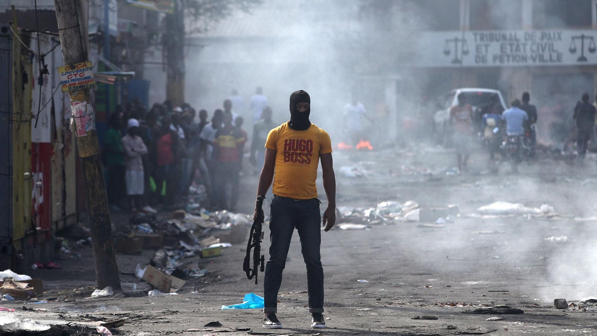 A man is shown holding a weapon, wearing a yellow Hugo Boss t-shirt, next to burning barricades.