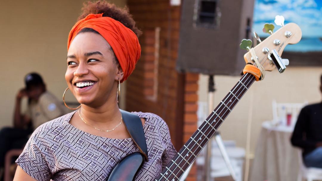 A woman wearing a colorful headband holds a bass guitar