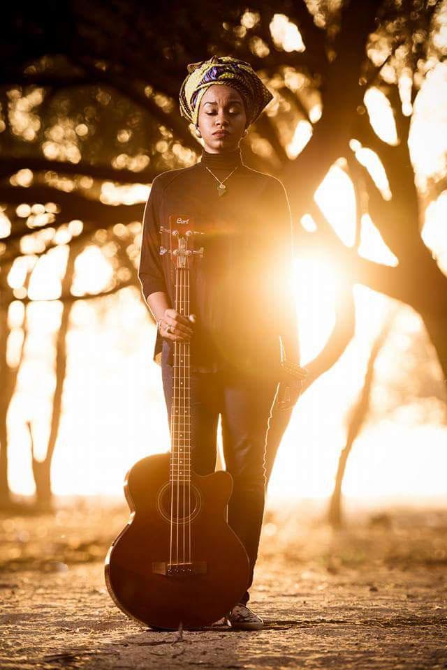 A woman poses with the sun behind her with a bass guitar