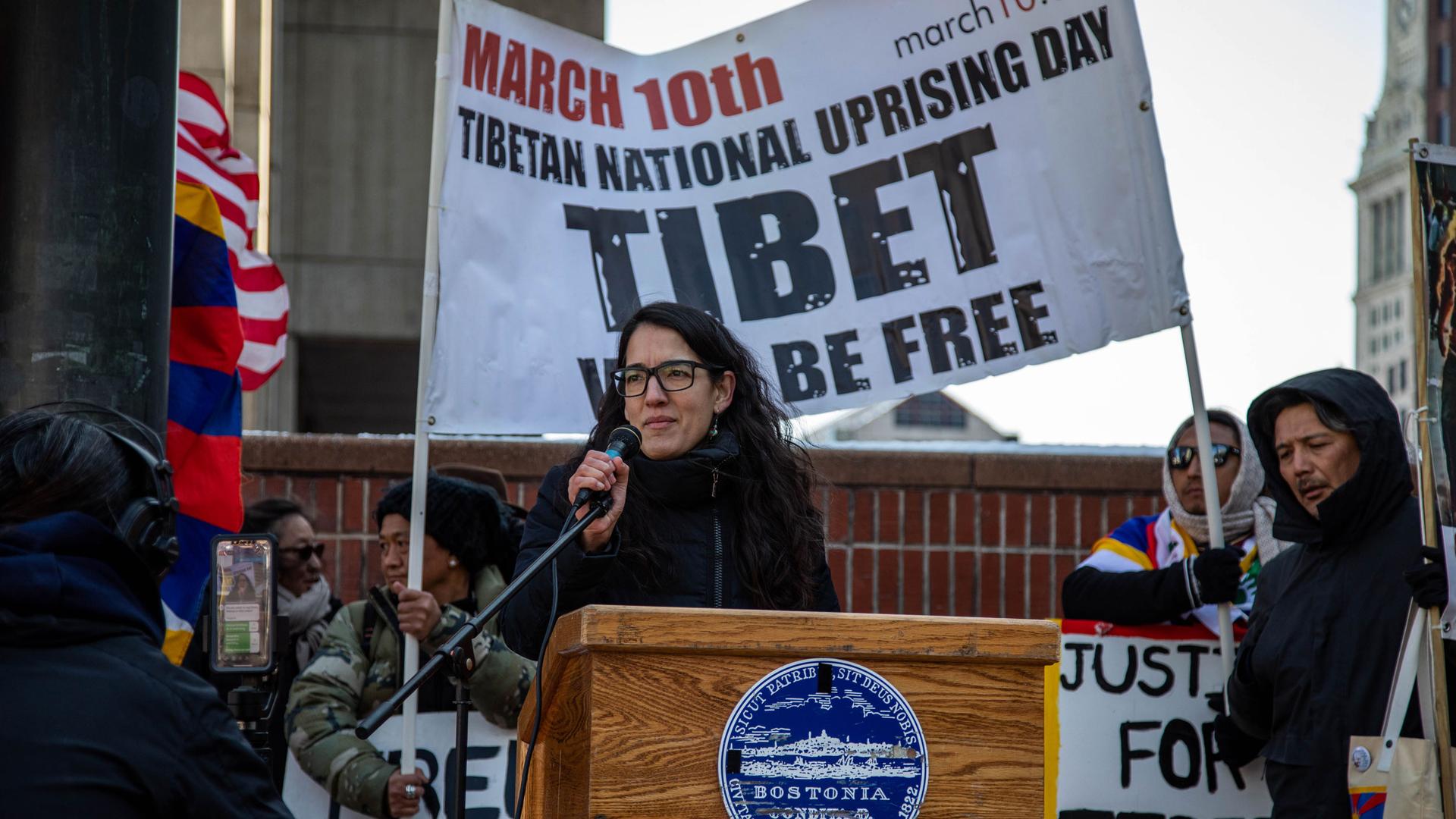 A woman stands at a podium. Behind her is a sign that says "Tibet will be free."