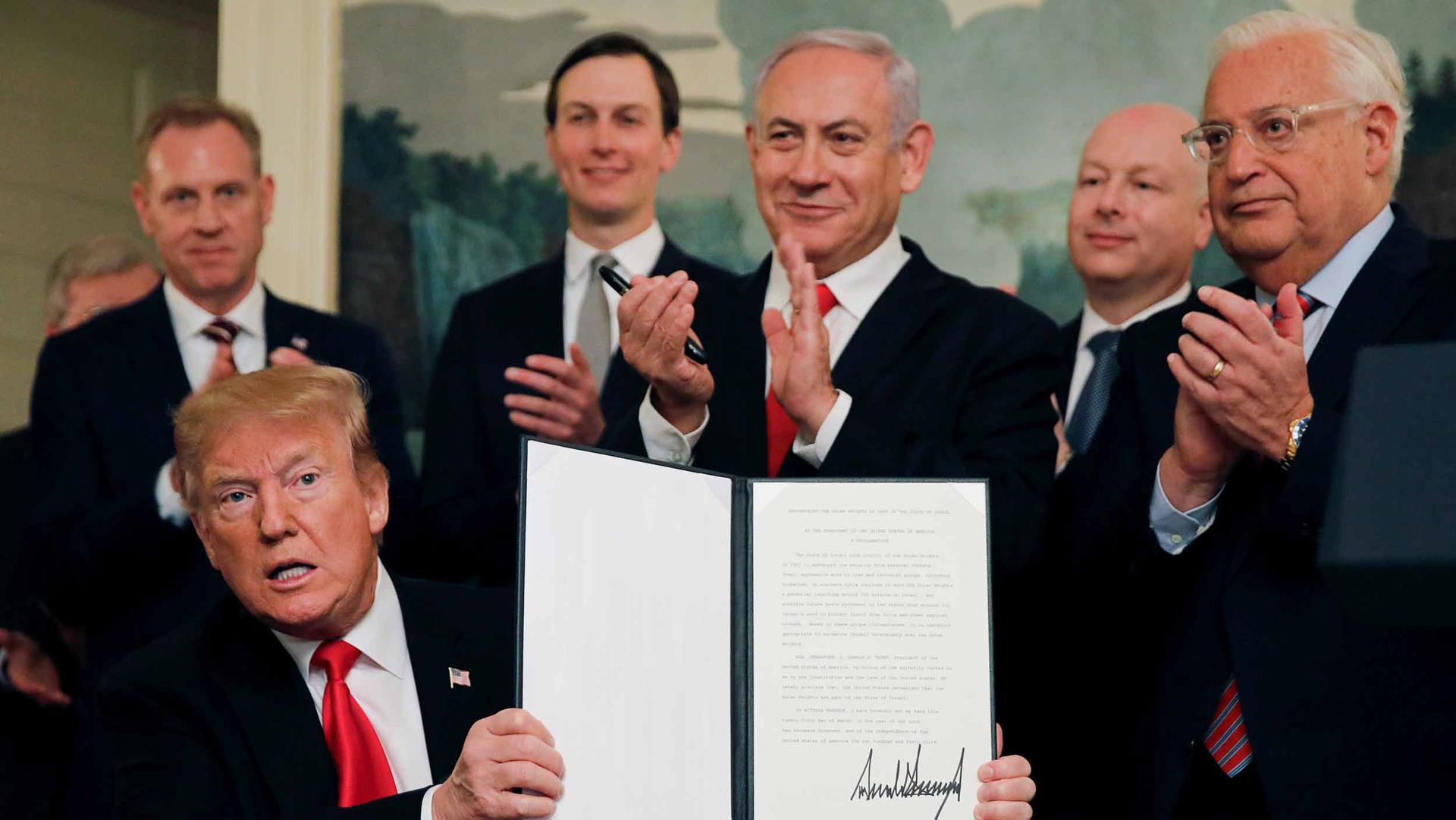 Donald Trump sits and holds up a signed proclamation while flanked by Israeli Prime Minister Benjamin Netanyahu and Jared Kushner