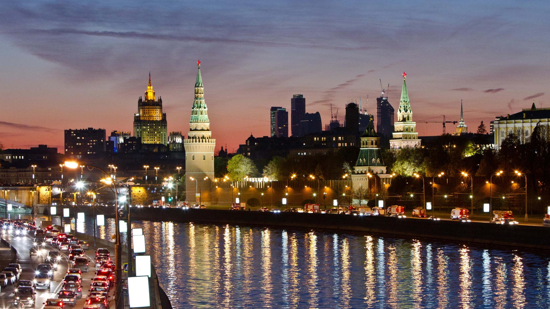 An evening cityscape of Moscow with towers in the background and a river and traffic-filled road in the foreground
