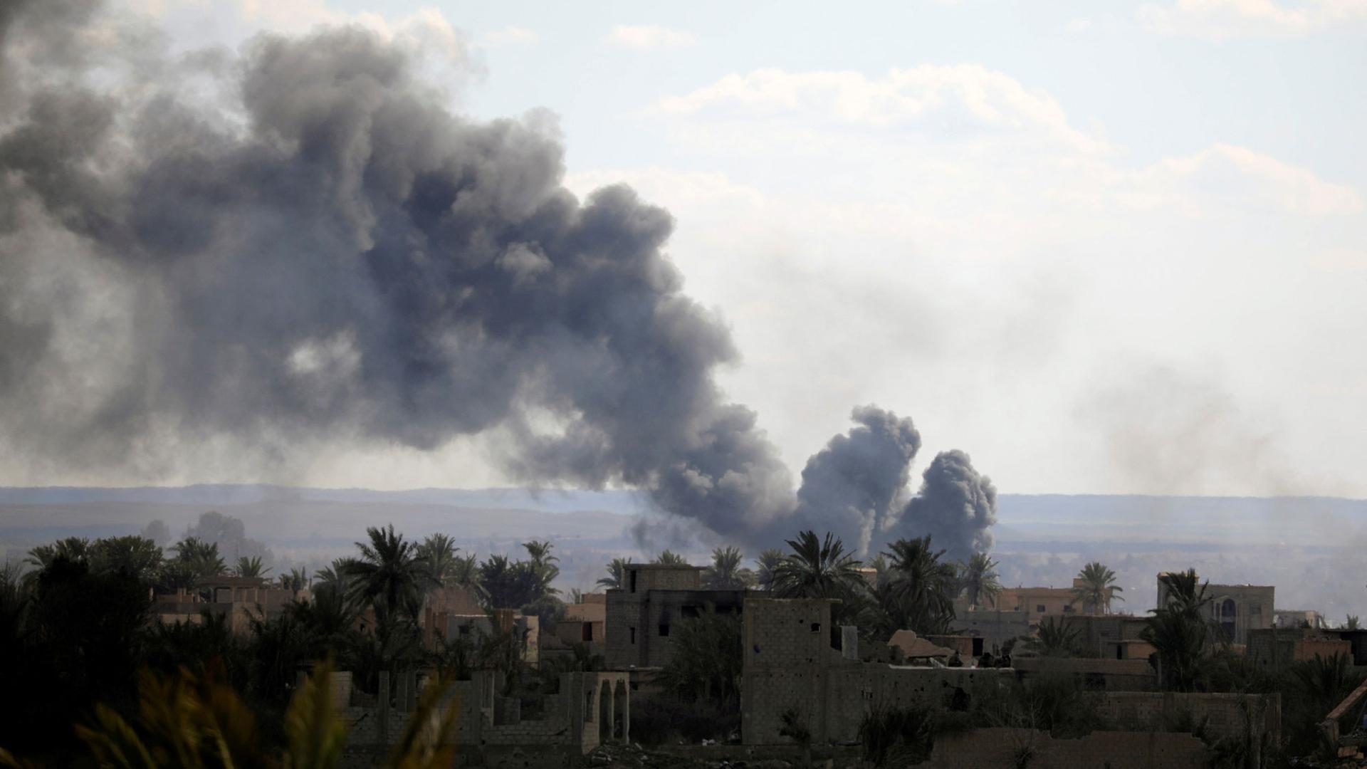 Black plumes of smoke are shown rising up and to the left of the photo with bombed out buildings below in Baghouz, Syria.