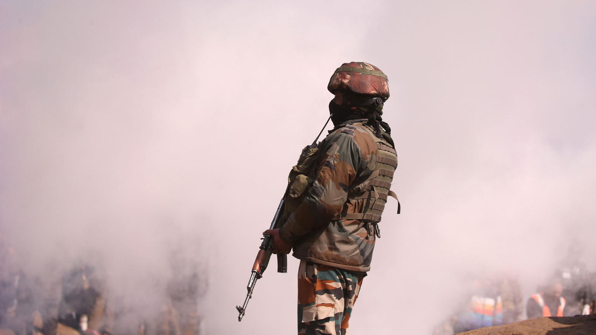 An Indian soldier stands guard while carrying a riffle with a lot of white smoke billowing behind him.