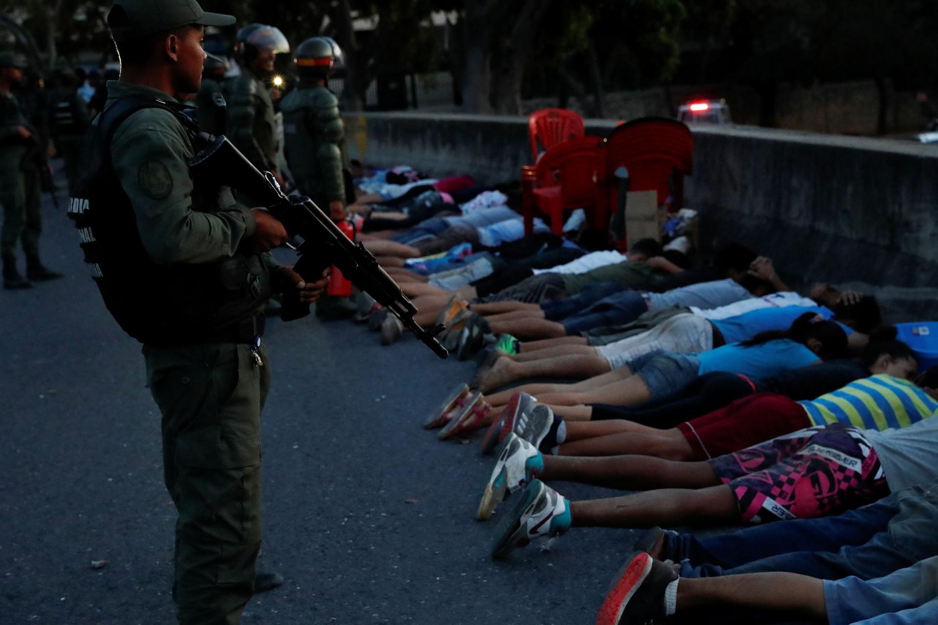 A security force member stands holding a rifle next to detainees laying on the street, face down.