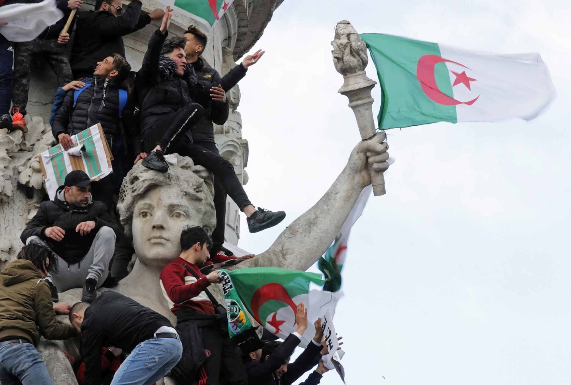 Many young men holding Algerian flags scale a monument of a woman holding a torch, attached to which is a flag