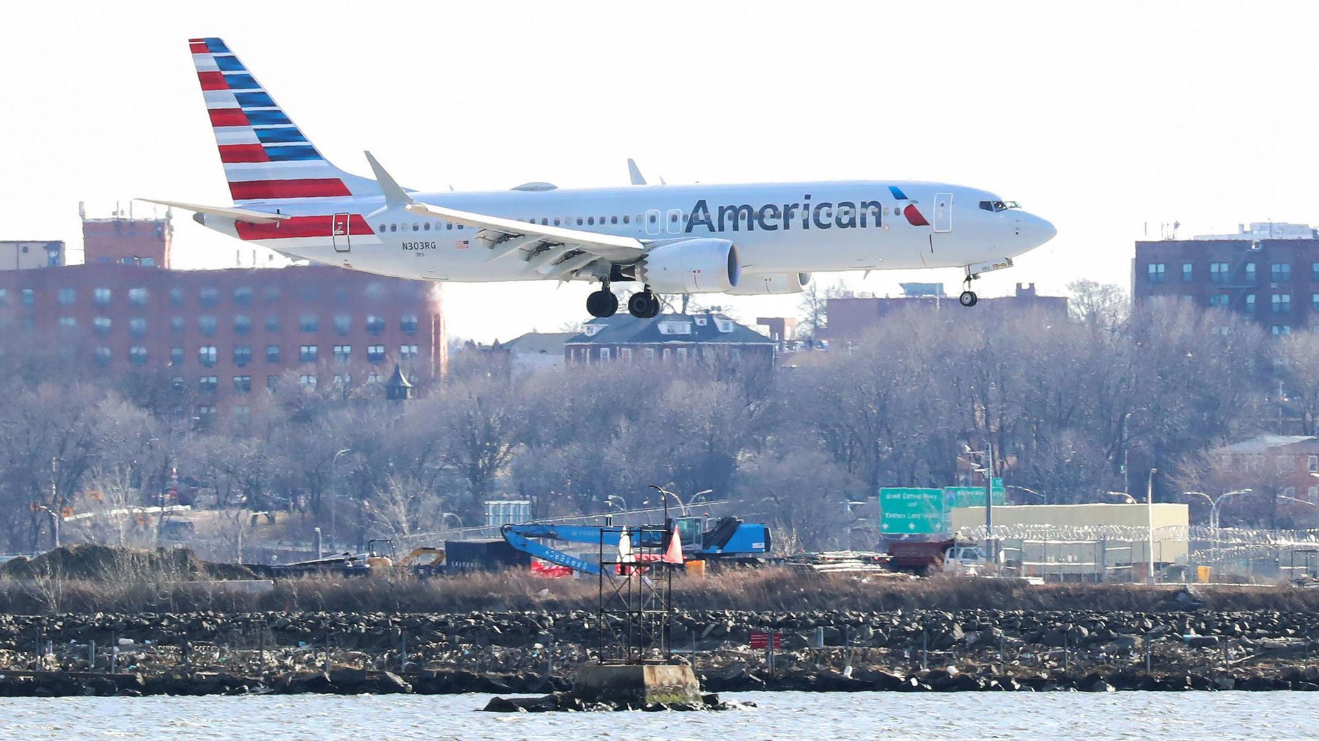 An American Airlines Boeing 737 Max 8 is shown flying from left to right in the photograph with several buildings in the background.