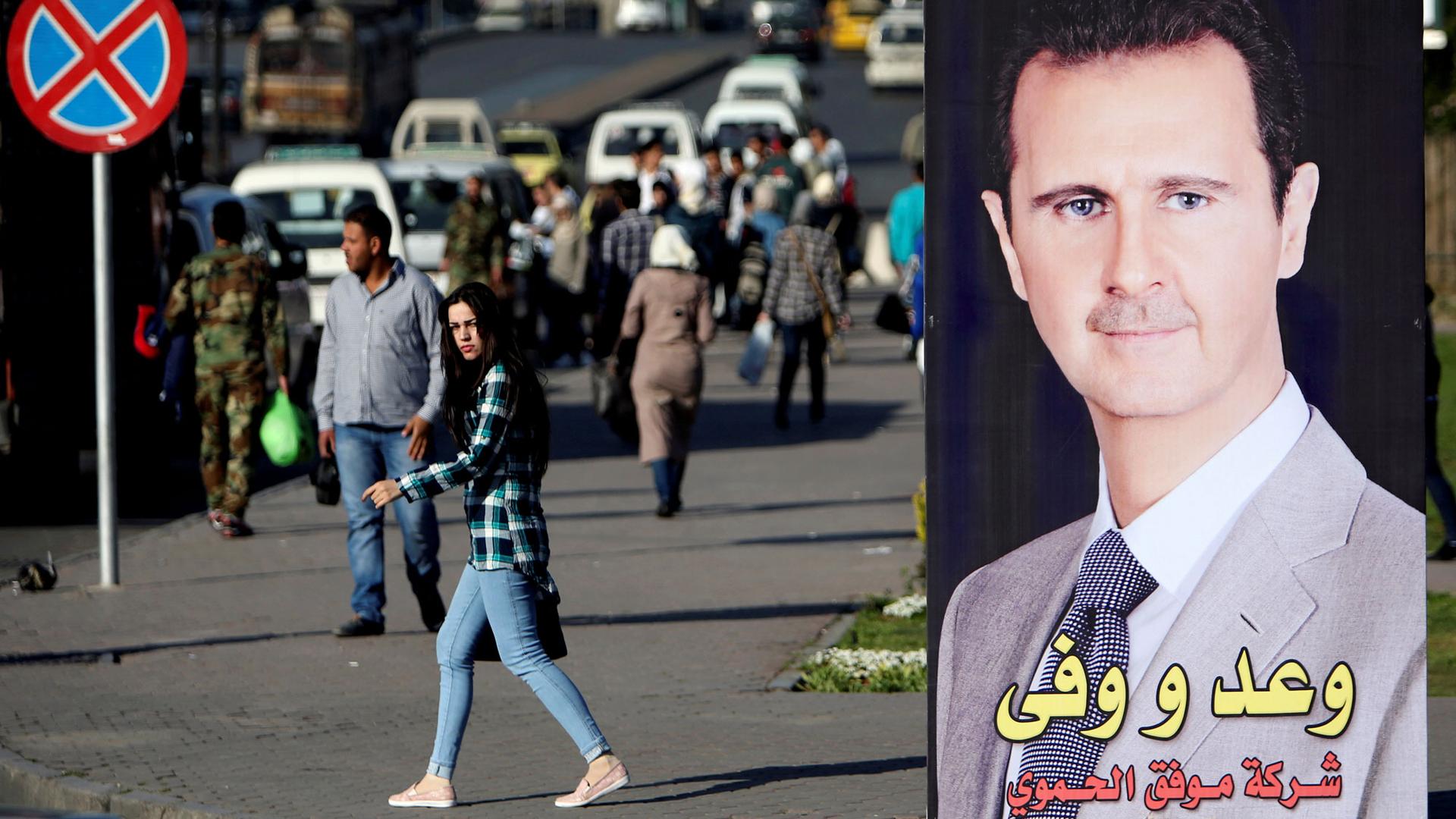A woman walks wearing jeans and a plaid shirt is shown near a picture of Syrian President Bashar al-Assad in Damascus.