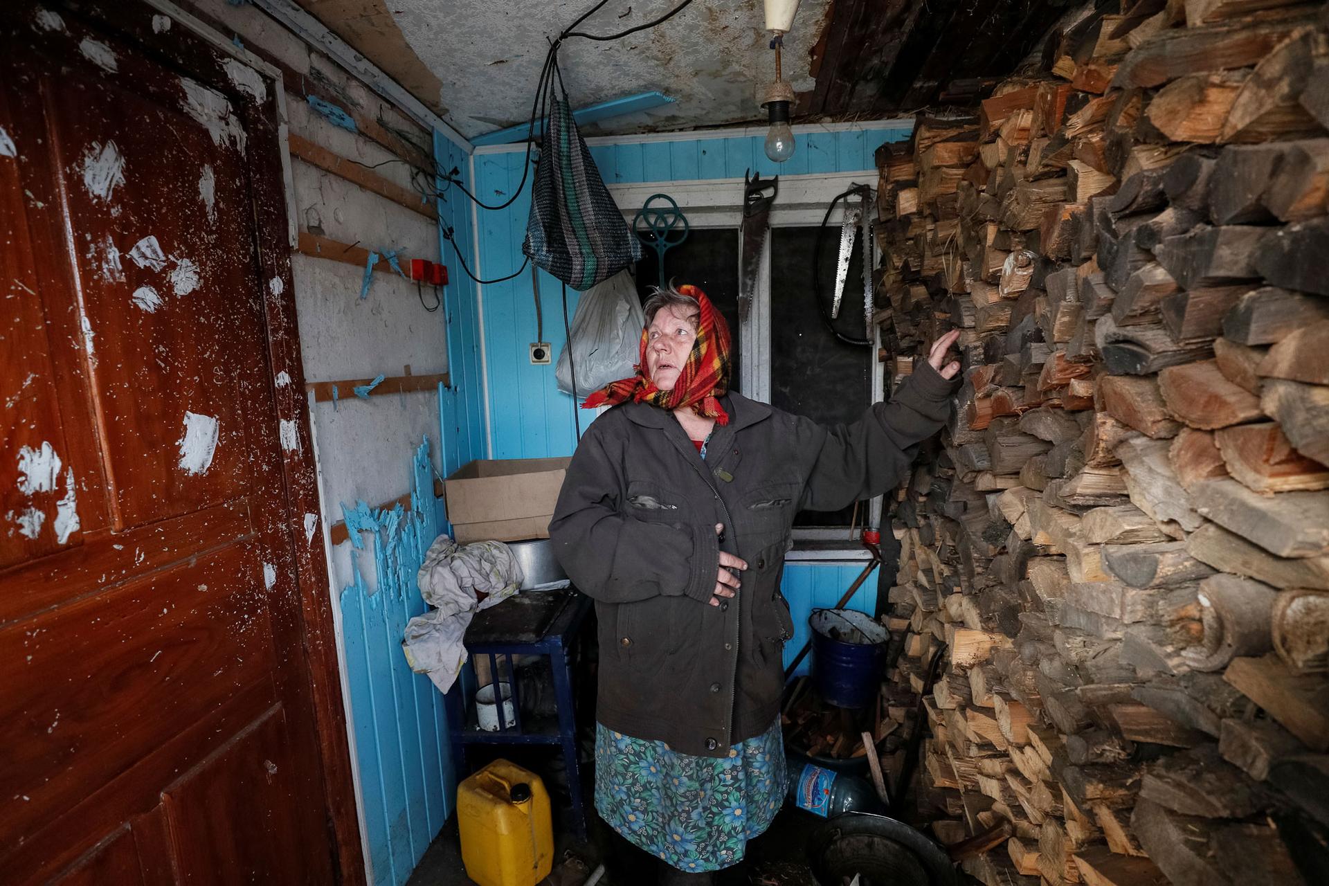A woman is shown with her hand on a wall of firewood looking to her right at bullet holes in door.