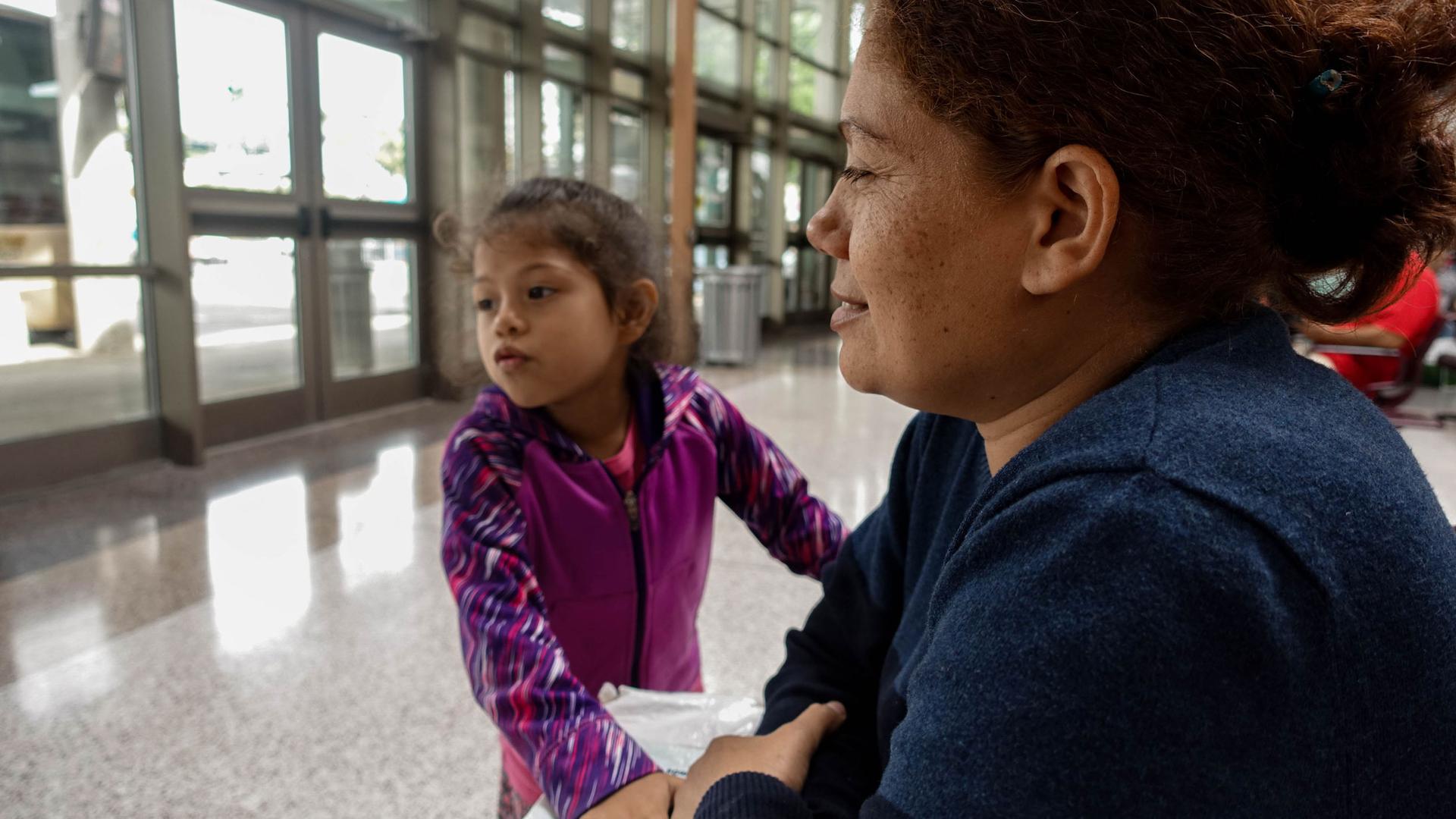 A close-up photo of a mother and her daughter, who is wearing pink, at a bus station. 