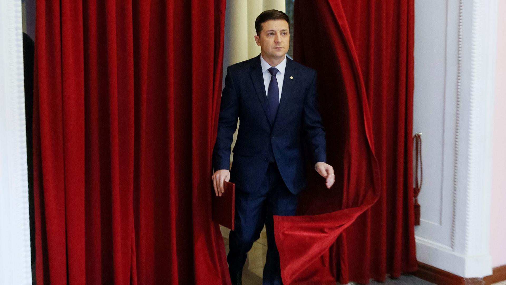 Actor Volodymyr Zelenskiy is shown in a blue suit walking out from parting red curtains.