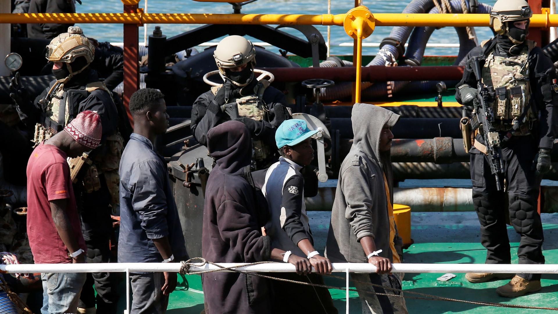 Migrants are shown in the near ground walking in a line as they disembark from a merchant ship with armed soldiers behind them.