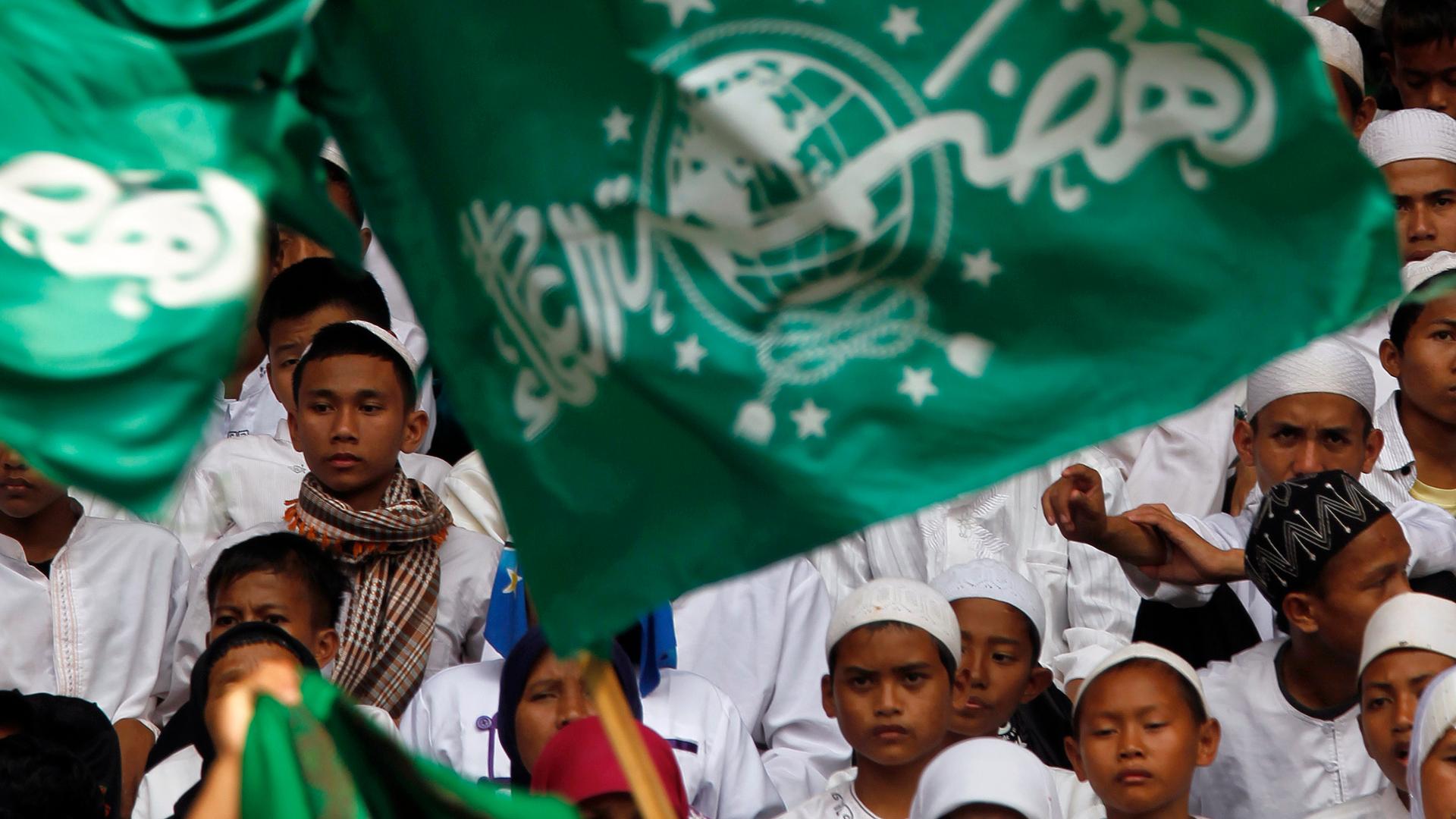 Young boys wearing white robes and caps stand behind green flag with Arabic script. 