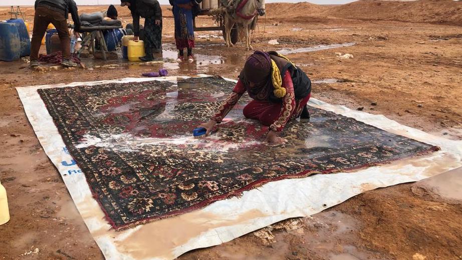 A woman crouches down and scrubs a large Persian rug in the desert. 