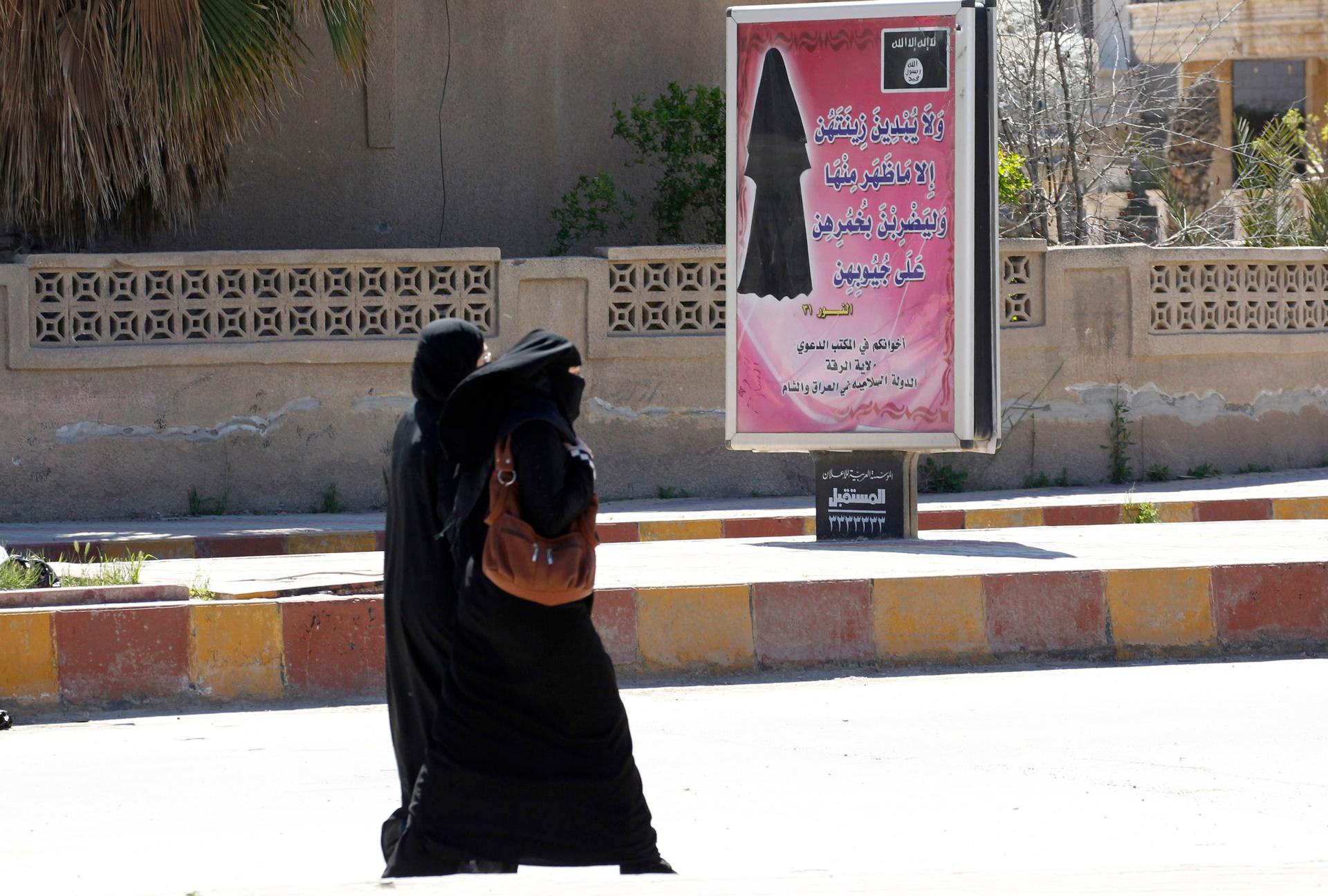 Two women wearing black veils walk outside past a sign with Arabic text. 