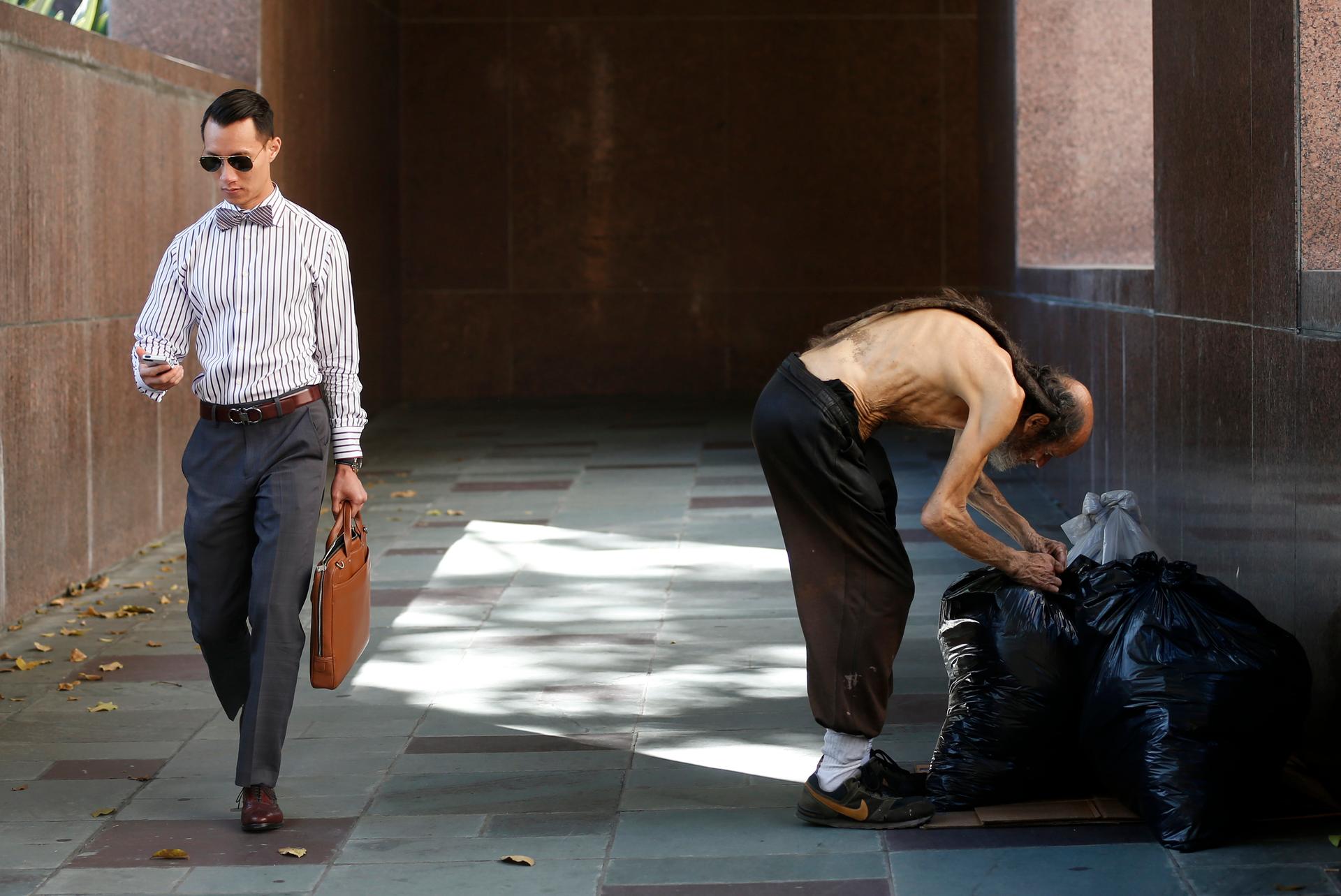 A man in business attire holding a briefcase walks past a shirtless man stooping over plastic bags