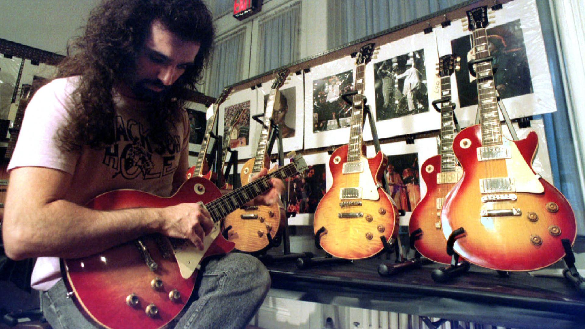 A man holds a guitar in his arms. Behind him are four more guitars on stands. 