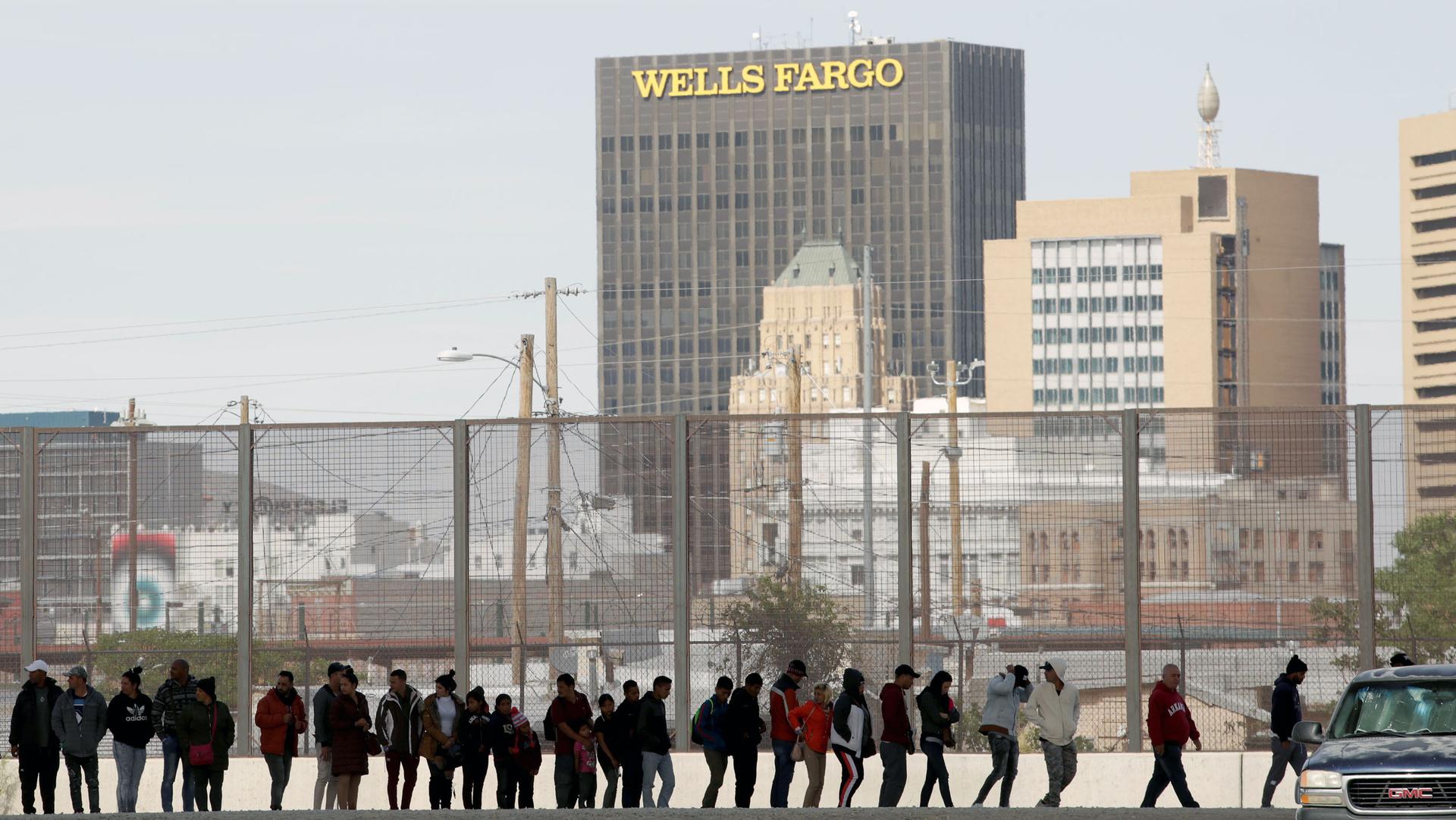 A line of people stand along a fence. Behind them, a high rise office tower reads "Wells Fargo"
