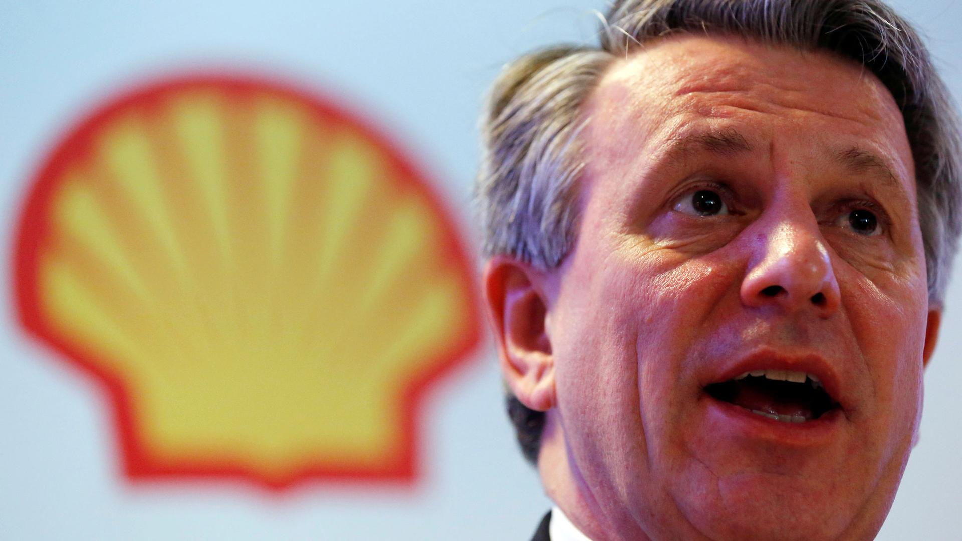 A man speaks with a Shell oil yellow shell logo behind him 