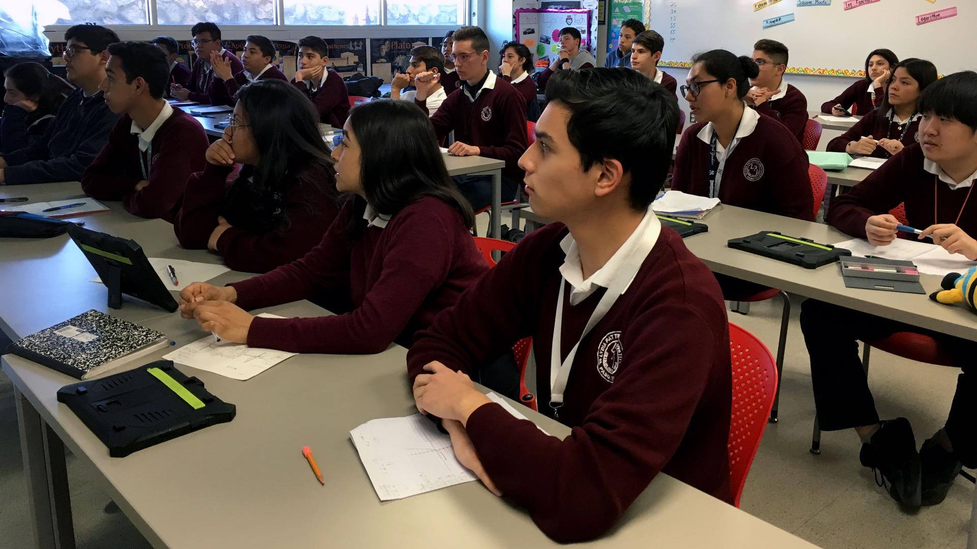 Students in maroon uniforms sit at desks and listen to a teacher, not pictured