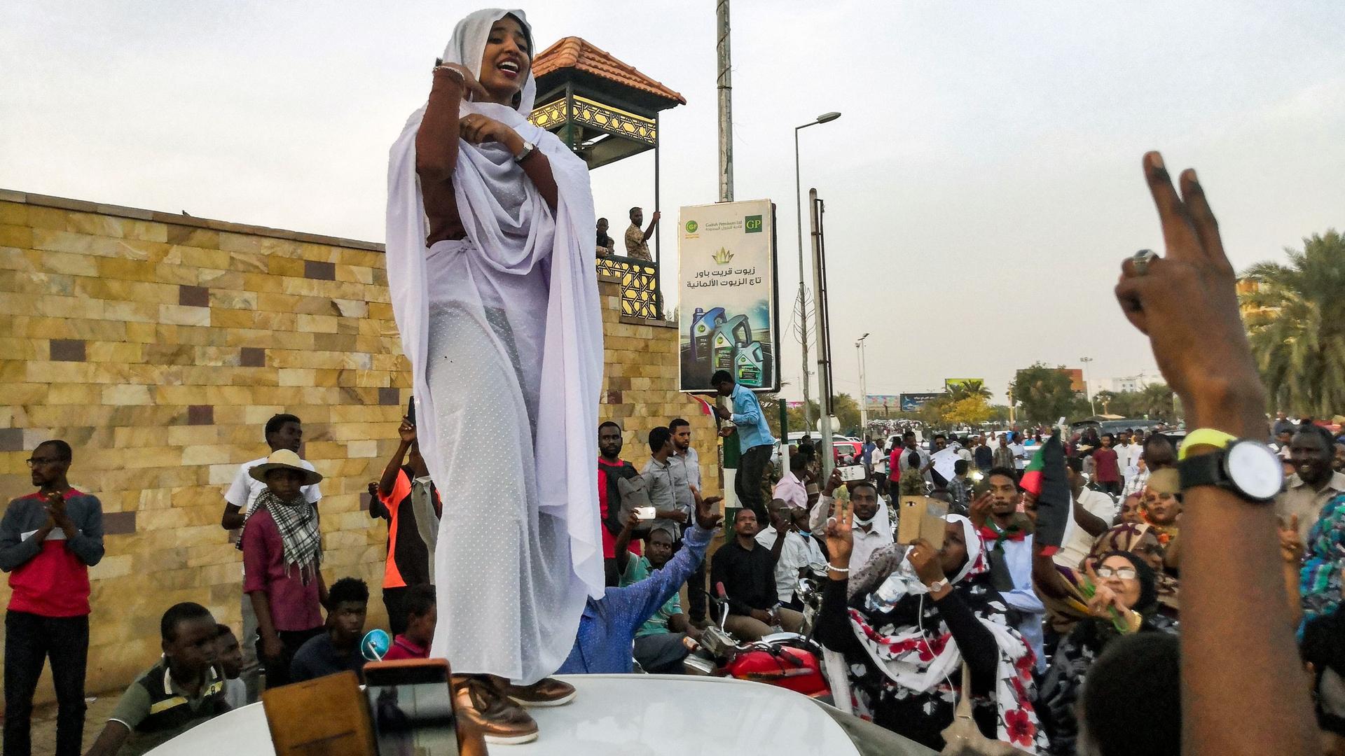 A woman stands atop a car and speaks to a crowd