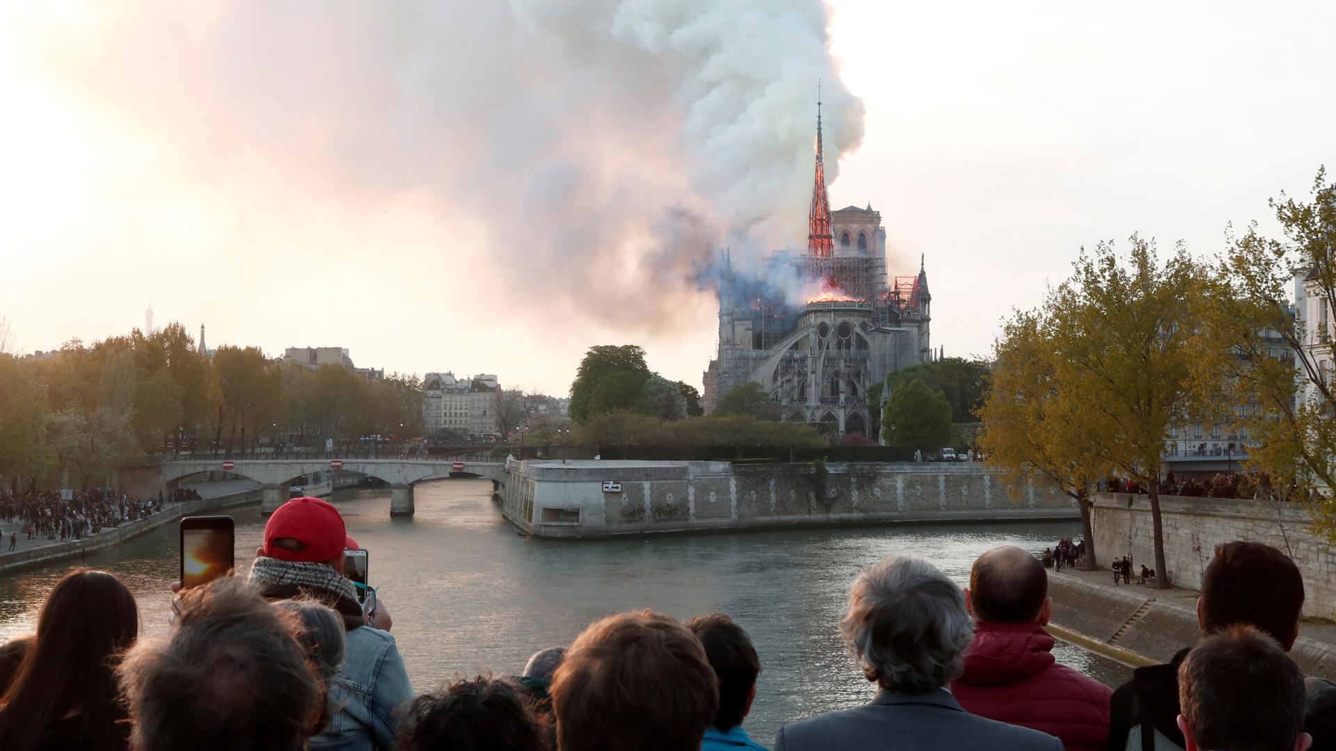 a crowd of people watch as the notre dame cathedral billows smoke in the distance