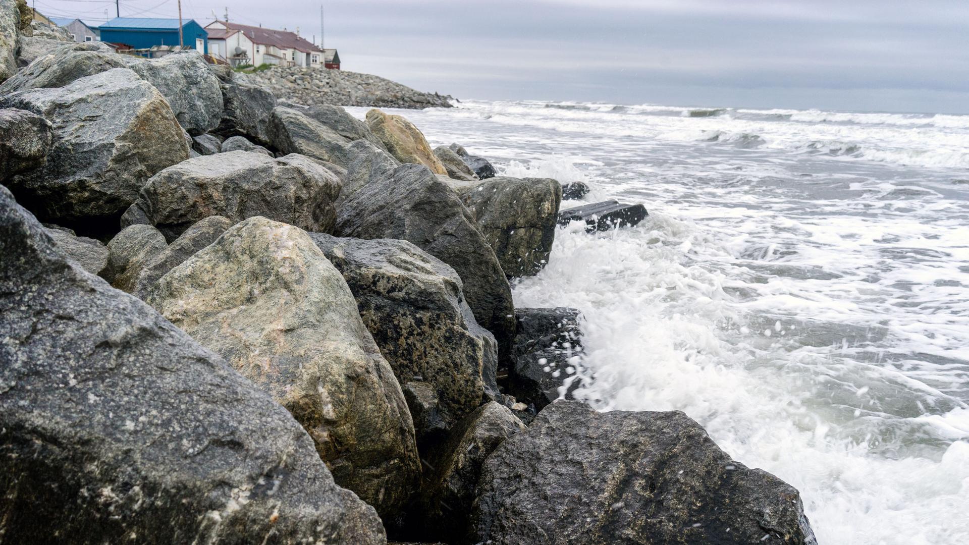 Waves crash into rocks. A house stands in the background.