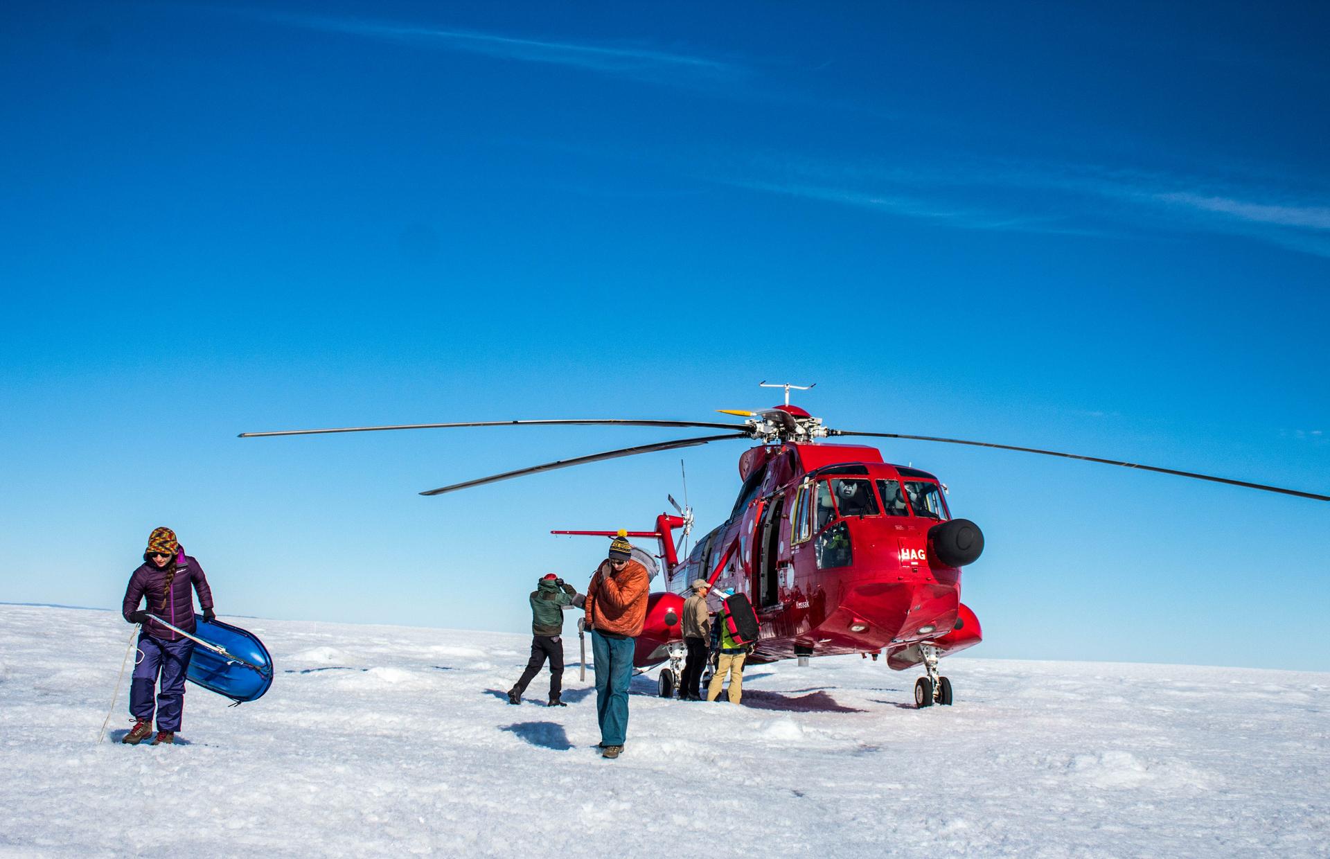 A red helicopter is shown landed on the Greenland ice sheet with team of three professors and three students unload their gear.