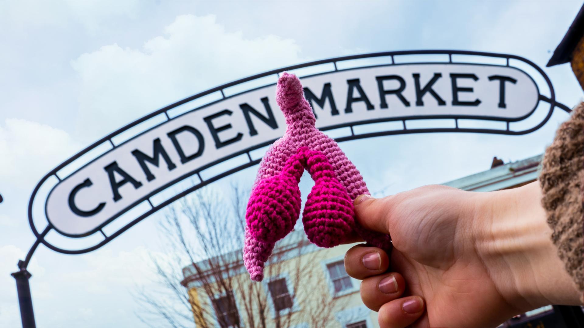 a knitted clitoris is held in front of the Camden Market sign 