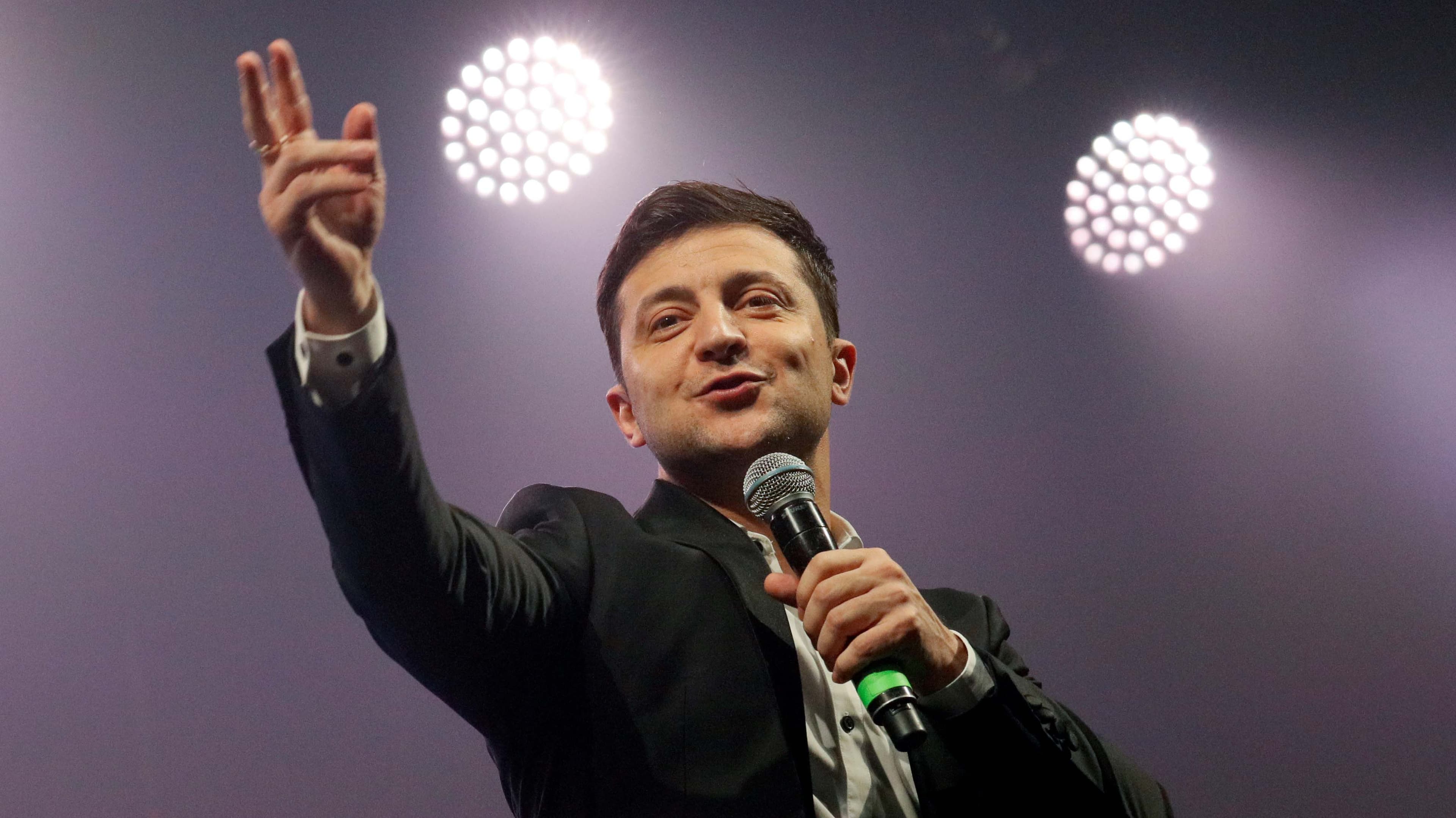 A man with a microphone in front of bright lights gives a peace sign.