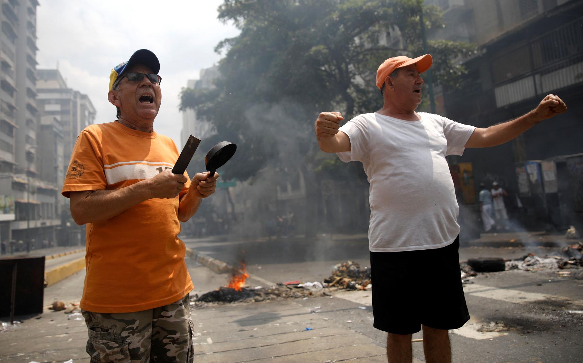A demonstrators wearing an orange shirt stand with a small pan in his hand next to another protester in a white t-shirt and orange hat.
