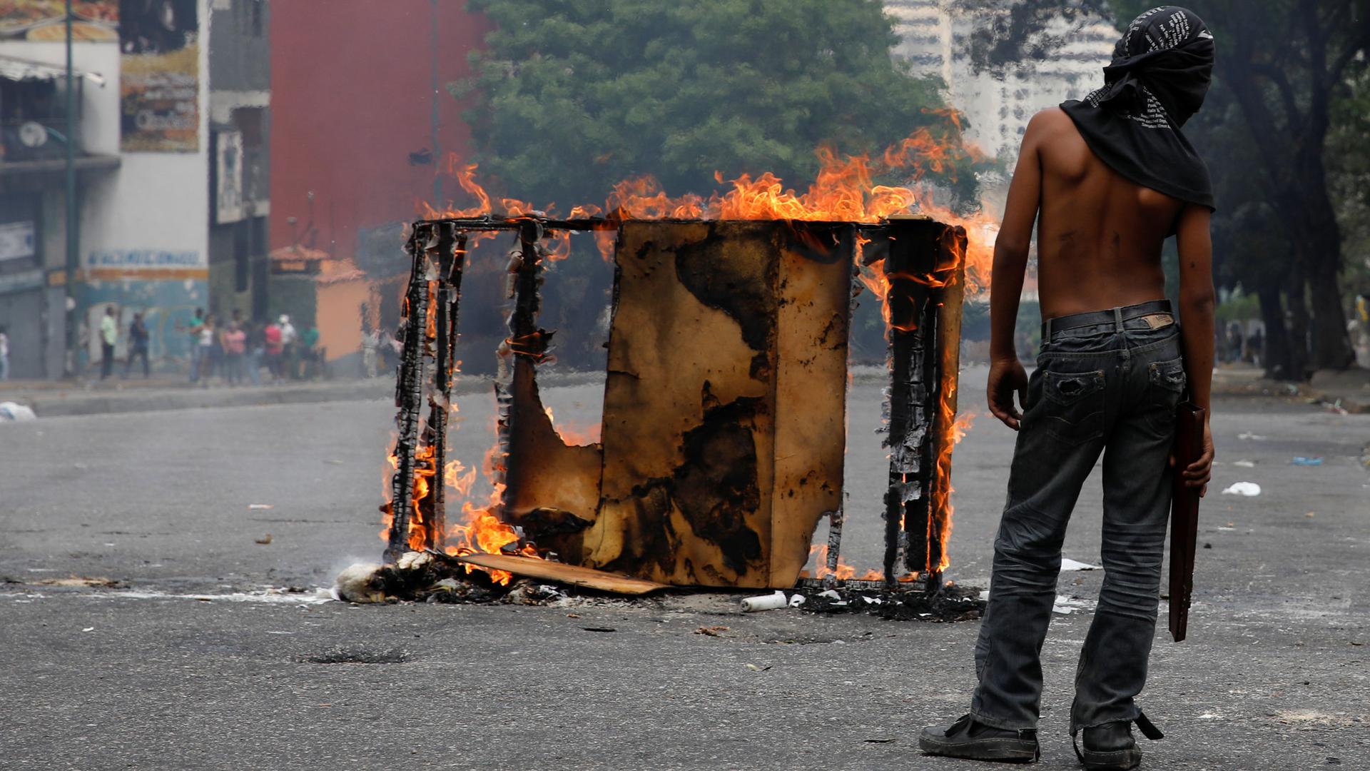 A demonstrator stands shirtless nex to a mostly burned rectangular frame during a protest .